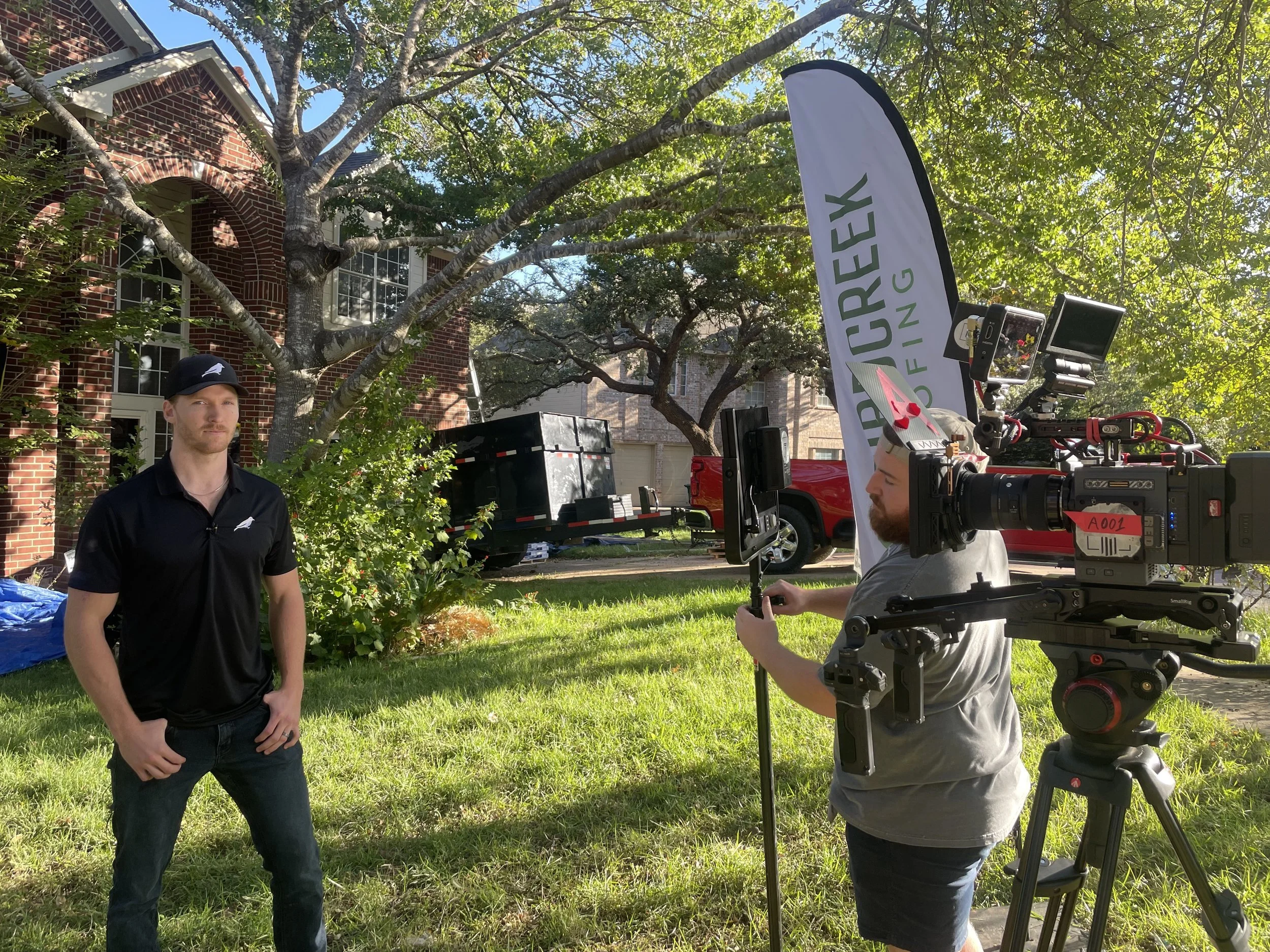 A man in black polo shirt and cap standing outdoors, being filmed by a camera operator with professional filming equipment, in front of a house with trees and a lawn.