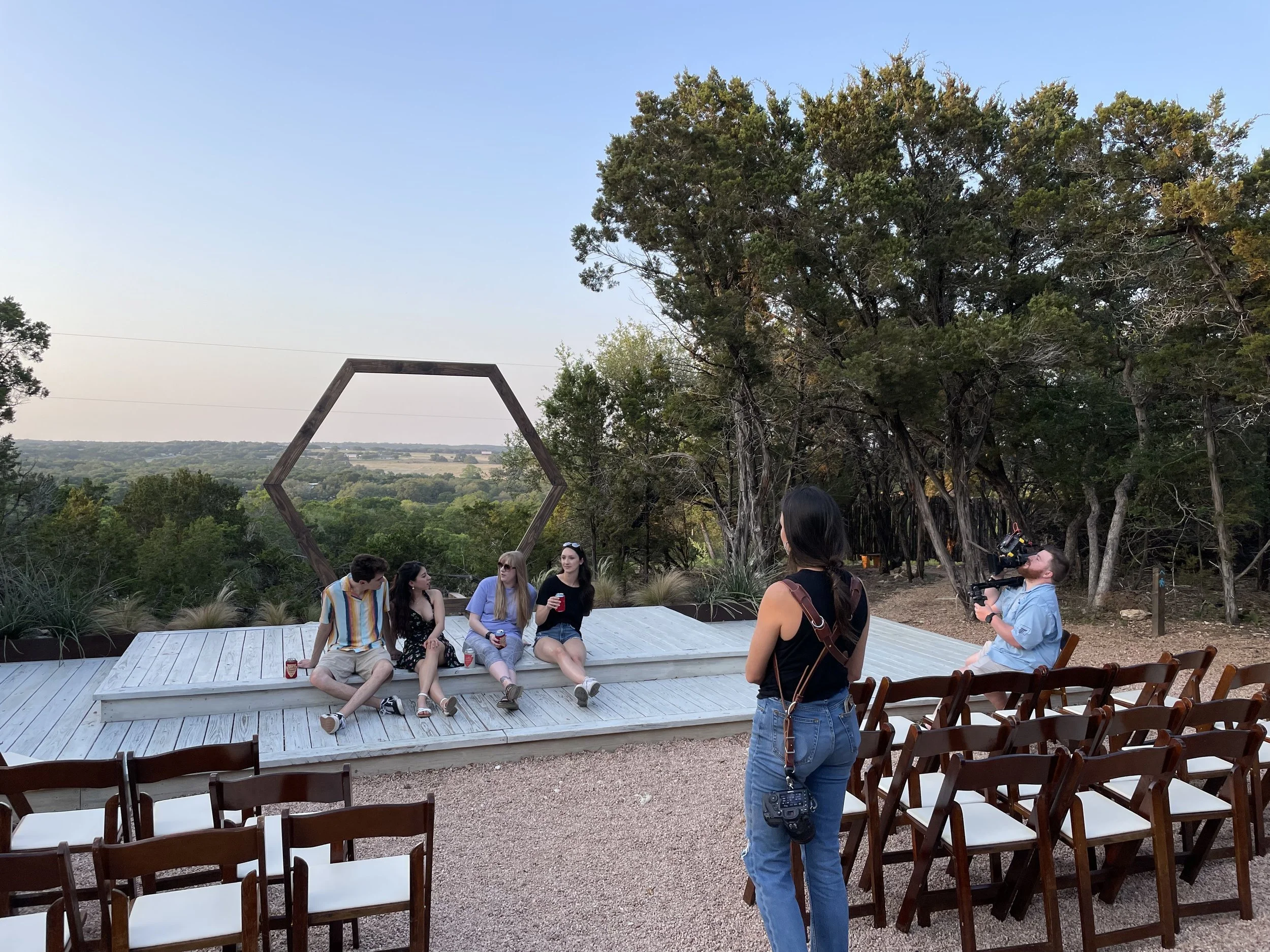 A group of four young women sitting on a white wooden platform outdoors, being filmed by a cameraman while a woman with a camera around her neck observes, in a wooded area with a hexagonal frame and distant landscape in the background.