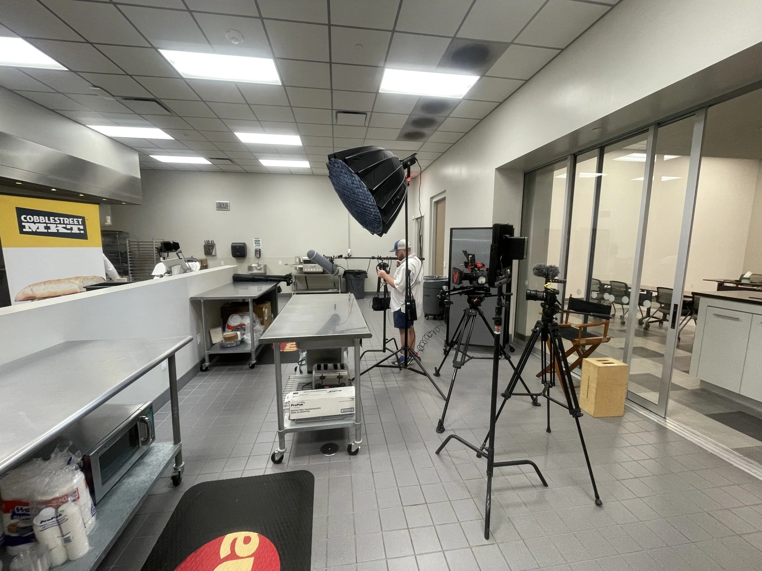 Behind the scenes setup in a commercial kitchen for filming, with camera equipment, lights, a person working with a camera, and a counter with kitchen supplies.