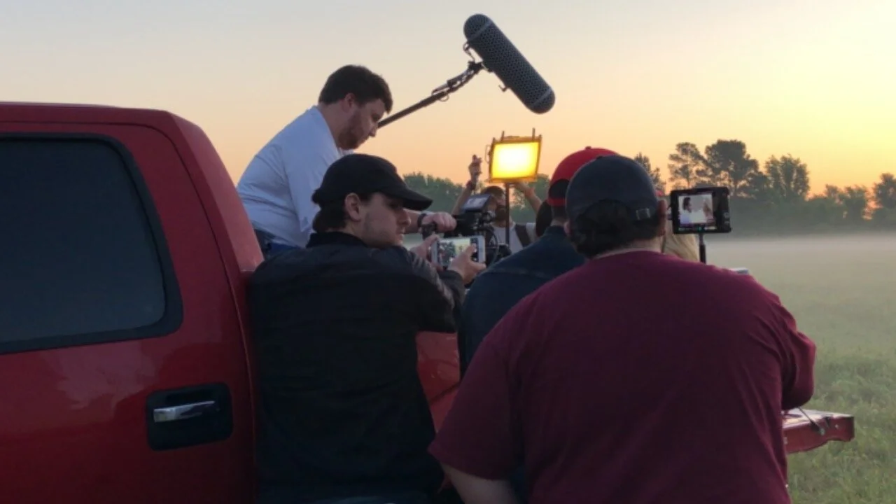 Filmmaking crew filming outdoors at sunrise or sunset, with equipment including a boom microphone and cameras mounted on tripods, on a grassy field with trees in the background.