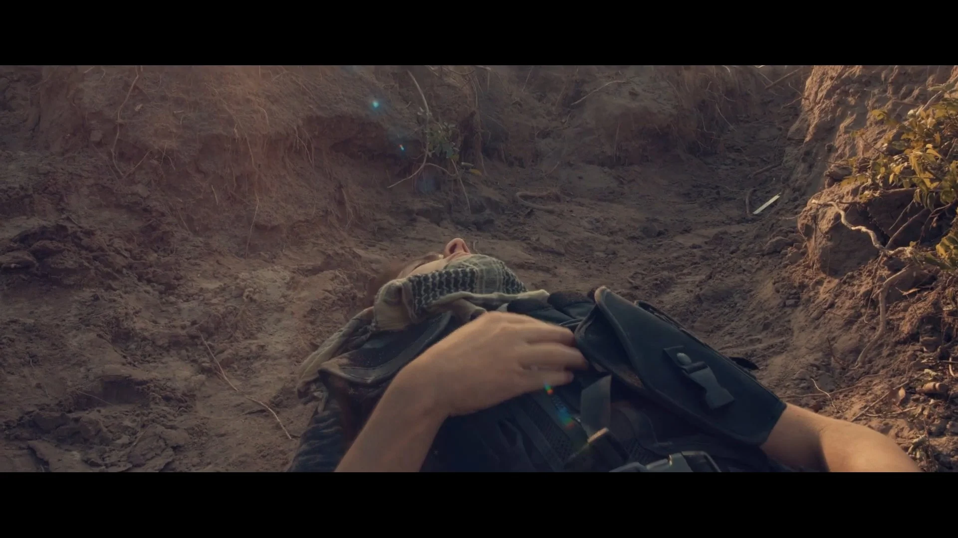 A person lying on the ground in a dirt trench, wearing a plaid scarf and tactical gear.