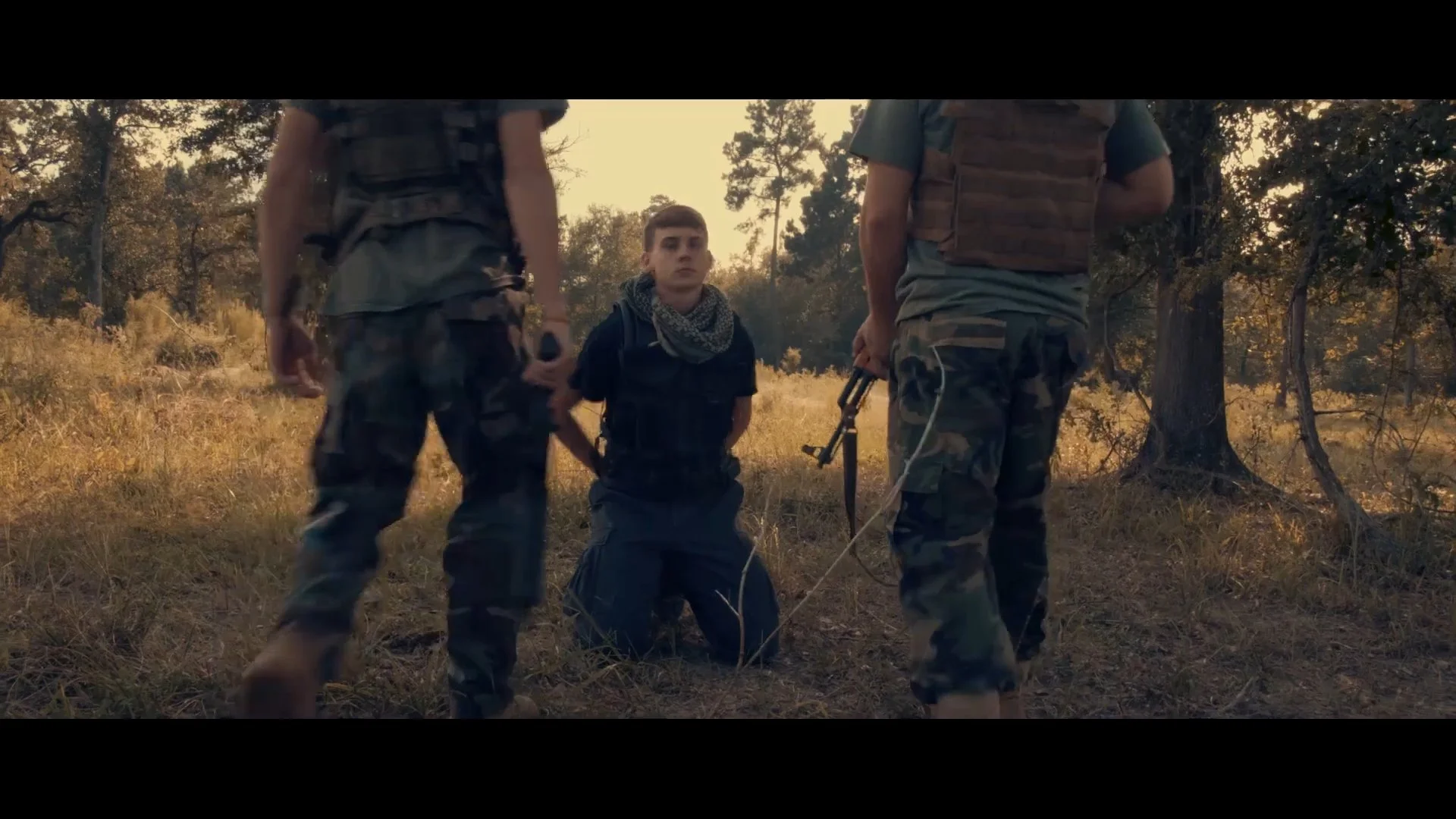 A person kneeling on the ground during an outdoor military or tactical operation with two others standing nearby, in a wooded area with trees and grass.