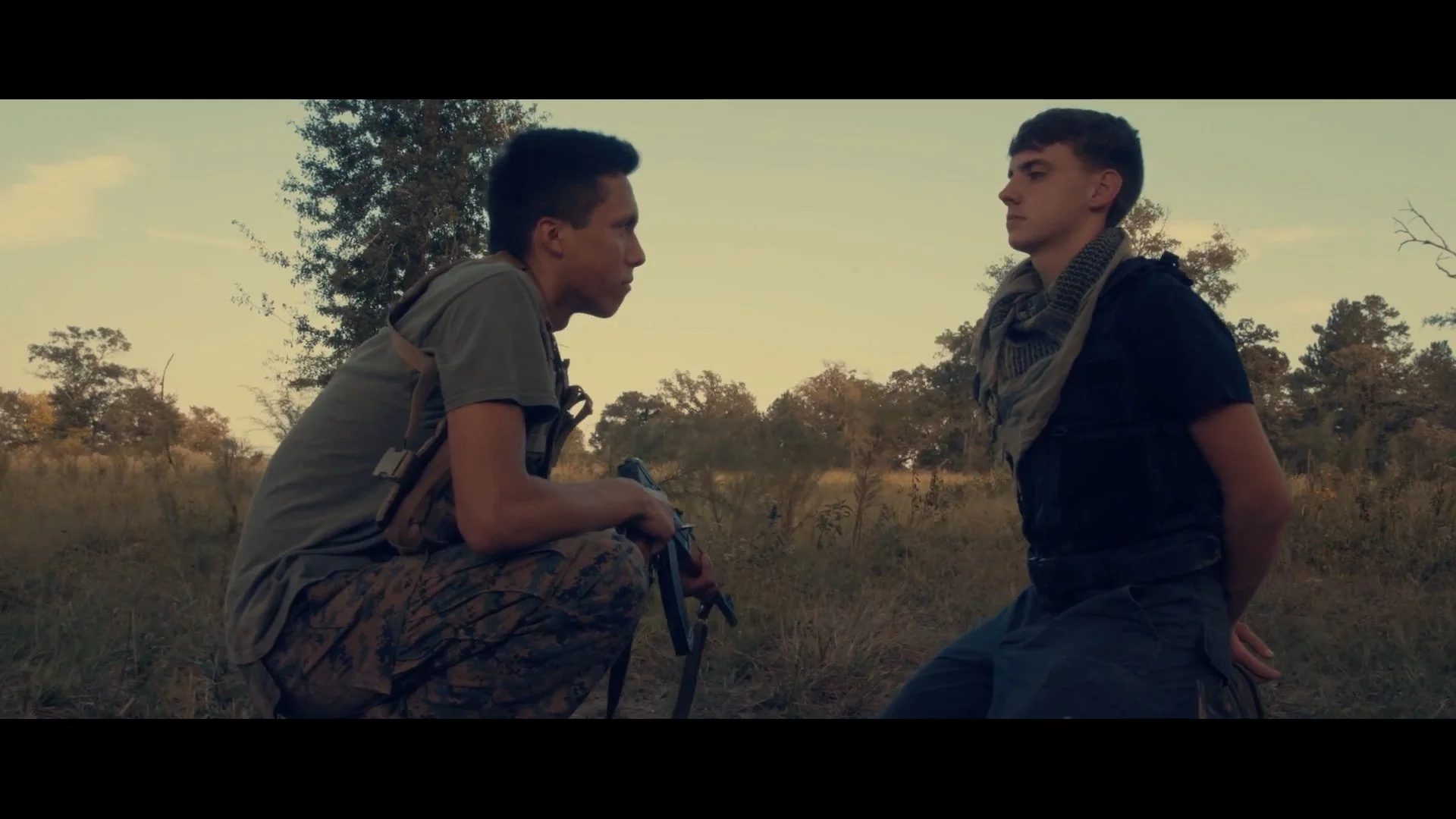 Two young men face each other in a grassy field, one kneeling with a backpack and the other standing with hands behind his back, in an outdoor setting with trees and a clear sky.