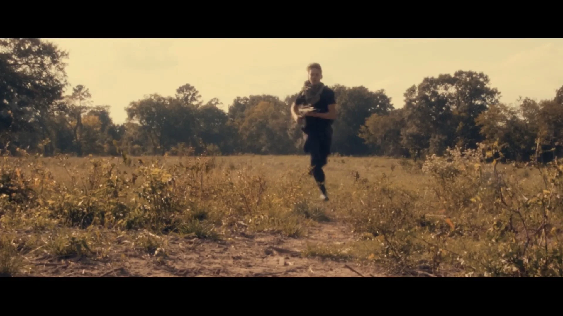 A person running through a field of plants with trees in the background on a sunny day.