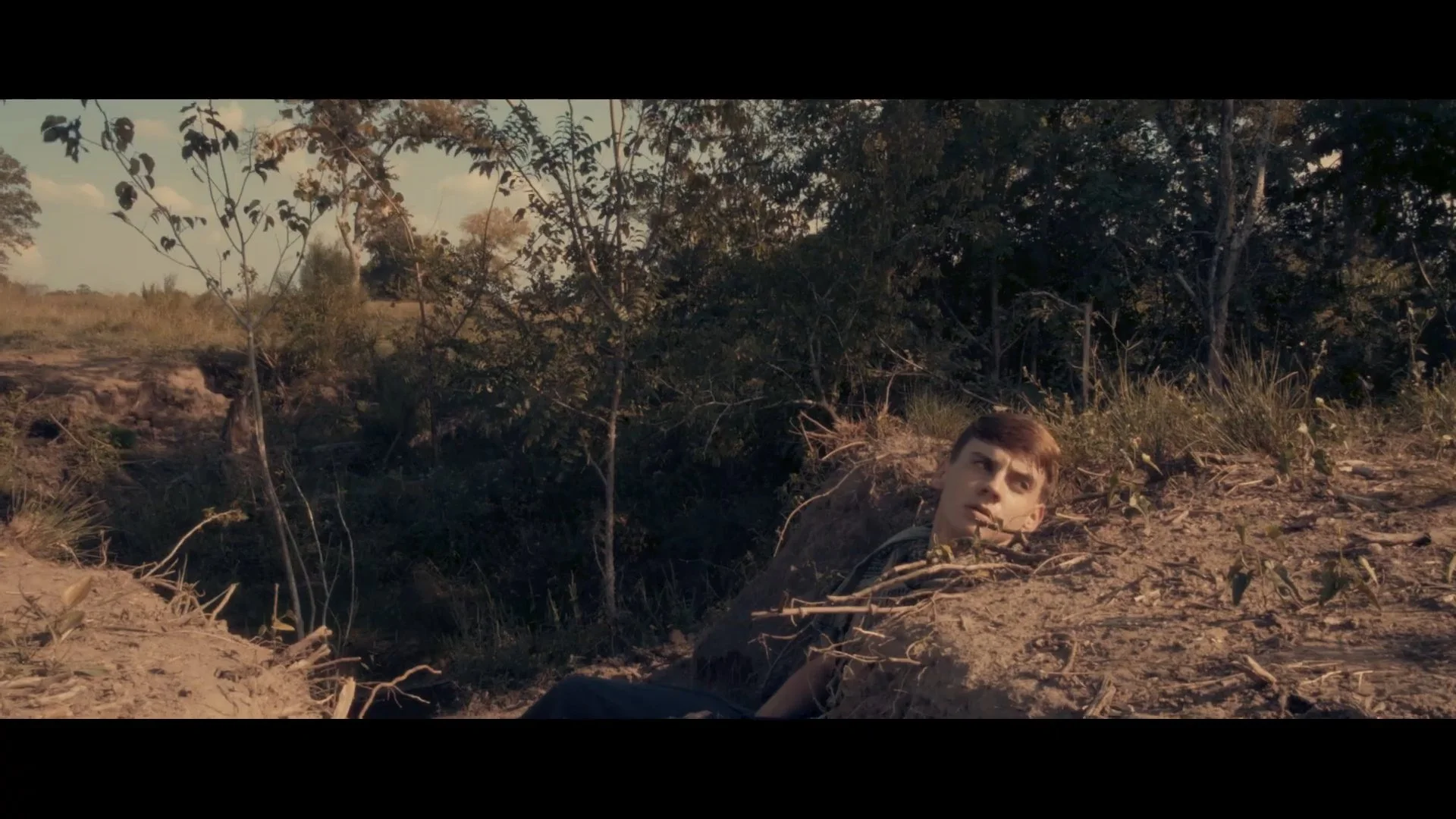 A young man lying on the ground in a dirt area surrounded by trees and bushes, looking alert and concerned.