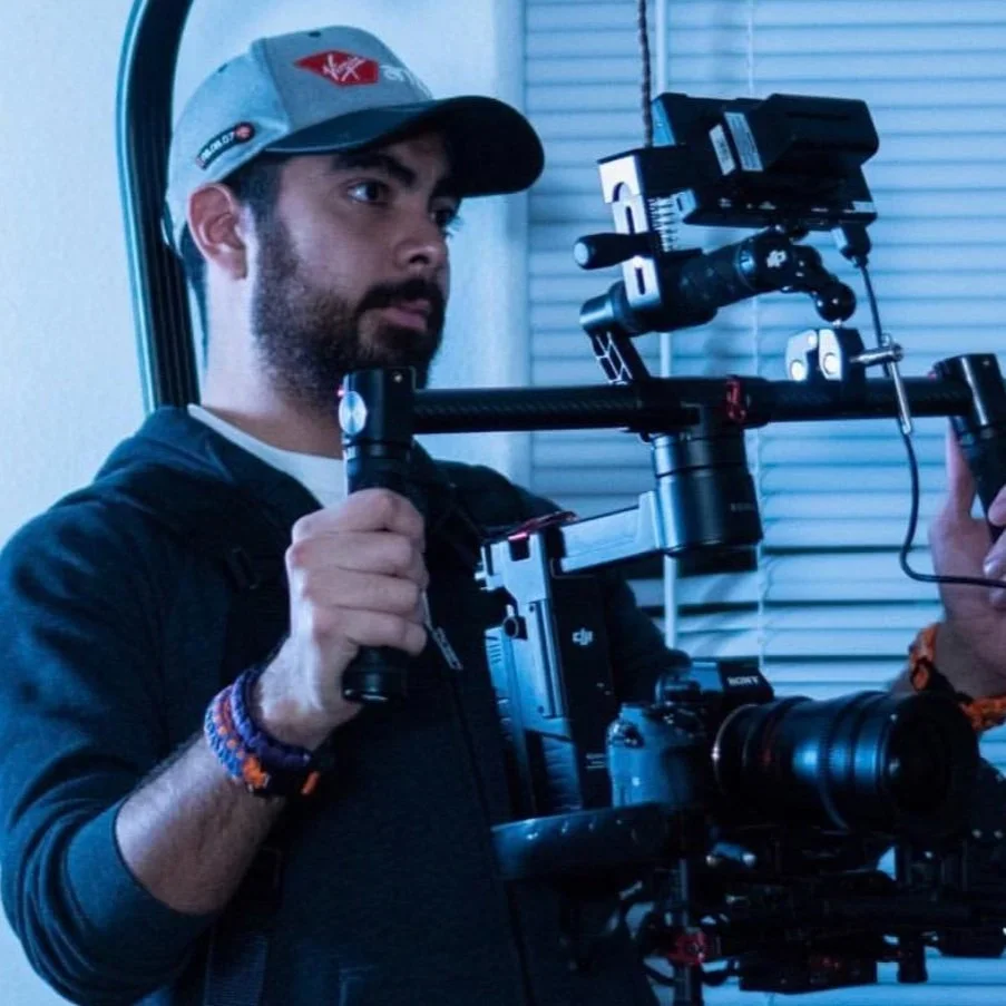 A man operating a professional camera stabilizer rig, wearing a baseball cap and dark clothing, indoors near a window with closed blinds.