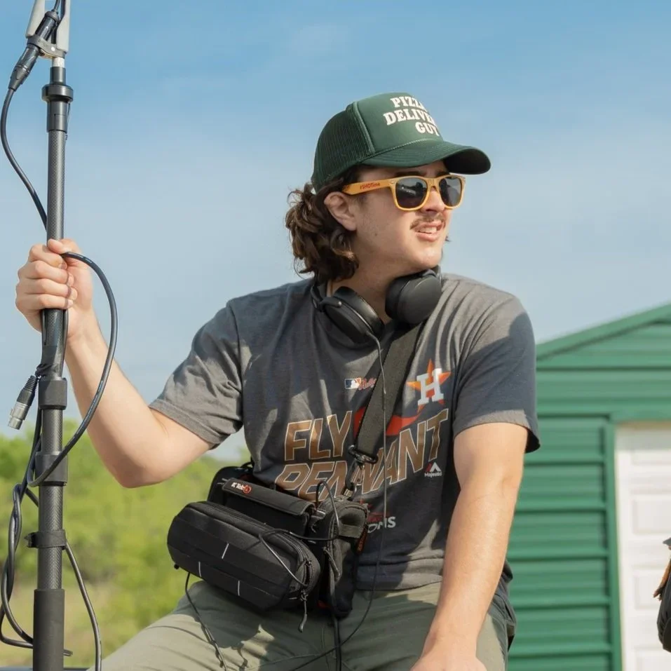 A man wearing a green cap, yellow sunglasses, and a gray T-shirt with orange and white lettering is holding a microphone stand outdoors on a clear day. He has headset headphones around his neck and a black camera bag hanging from his shoulder. He is looking off into the distance with a slight smile, with a green and white building in the background.