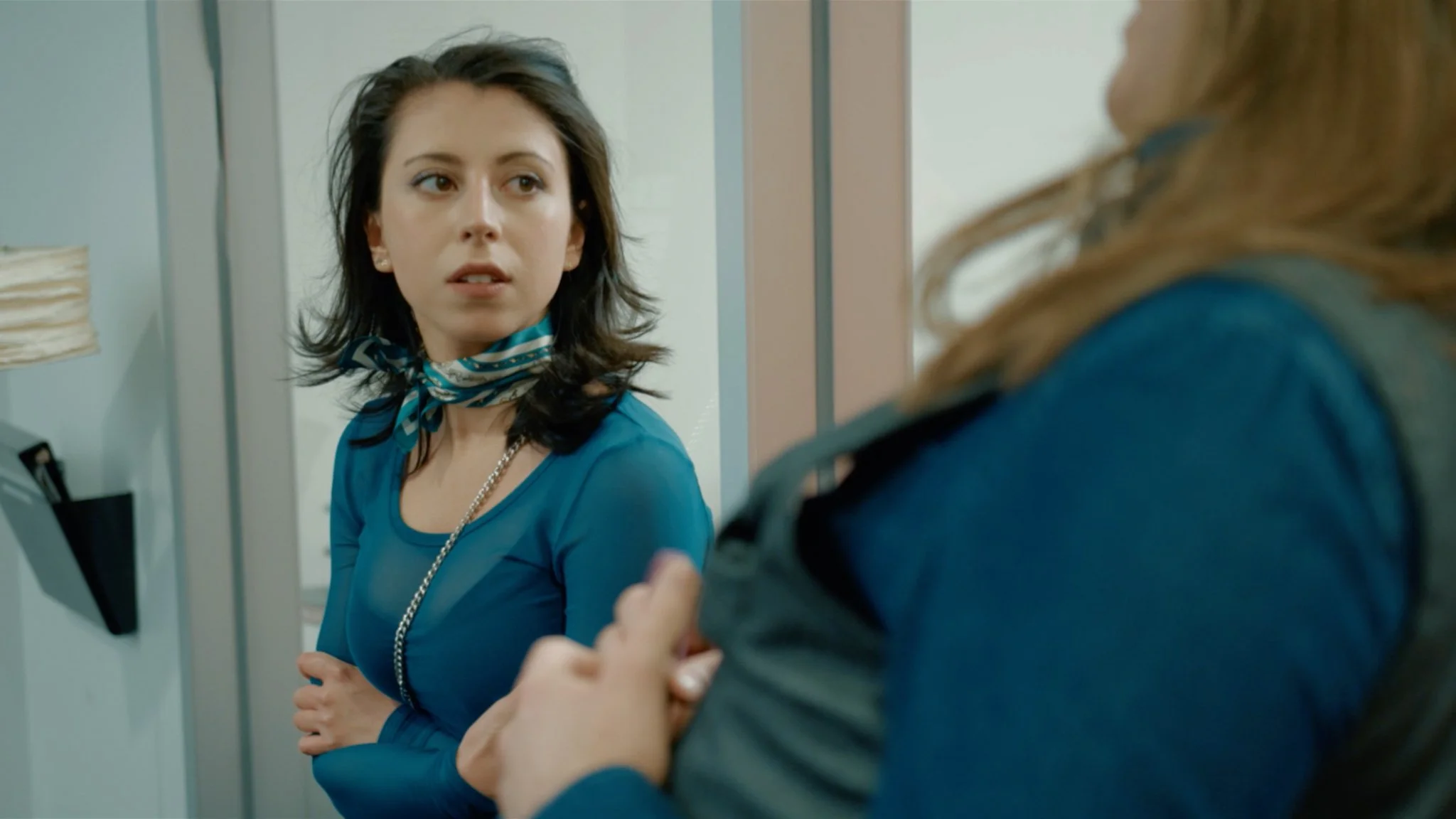 A young woman with dark hair, wearing a blue top and a scarf, looks surprised or concerned while talking to another woman who has her arm crossed and is partially out of the frame. They are in a room with light-colored walls and a telephone on the wa