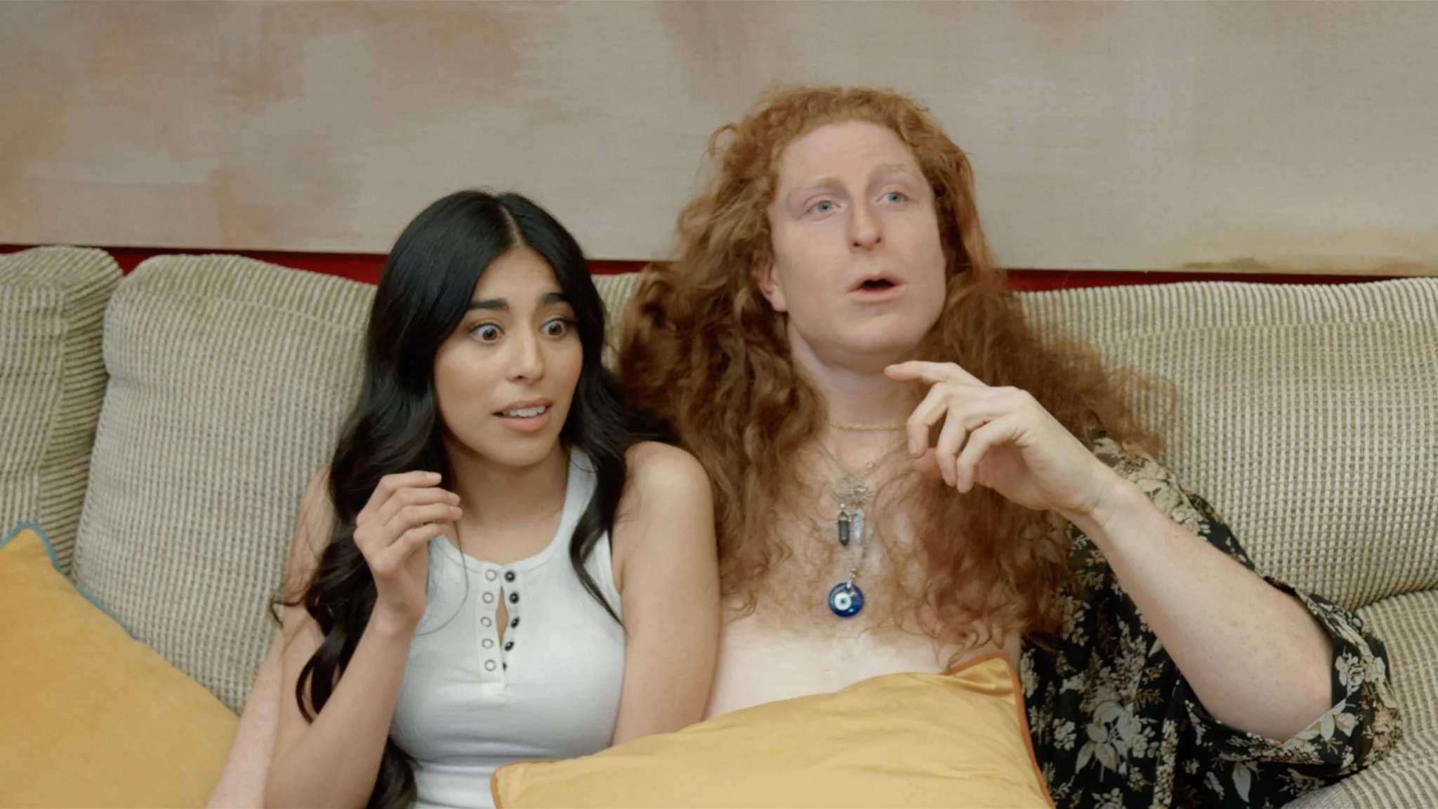 Two women sitting on a beige couch with surprised or shocked expressions, one with long black hair and the other with long curly red hair, wearing necklaces and relaxed clothing.