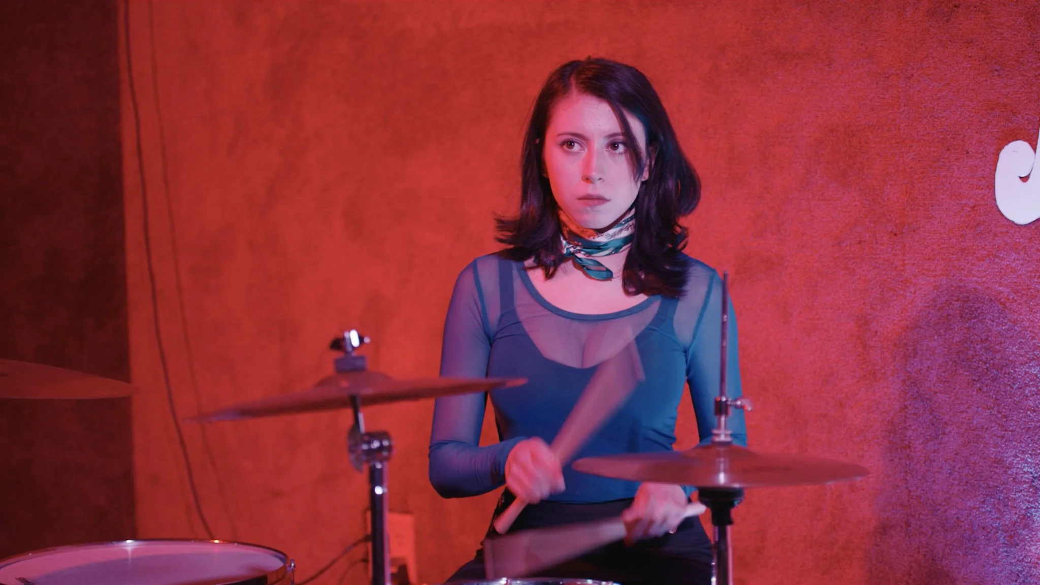 Young woman with dark hair wearing a semi-sheer blue top and a colorful scarf playing drums in a dimly lit room with a red-orange wall.