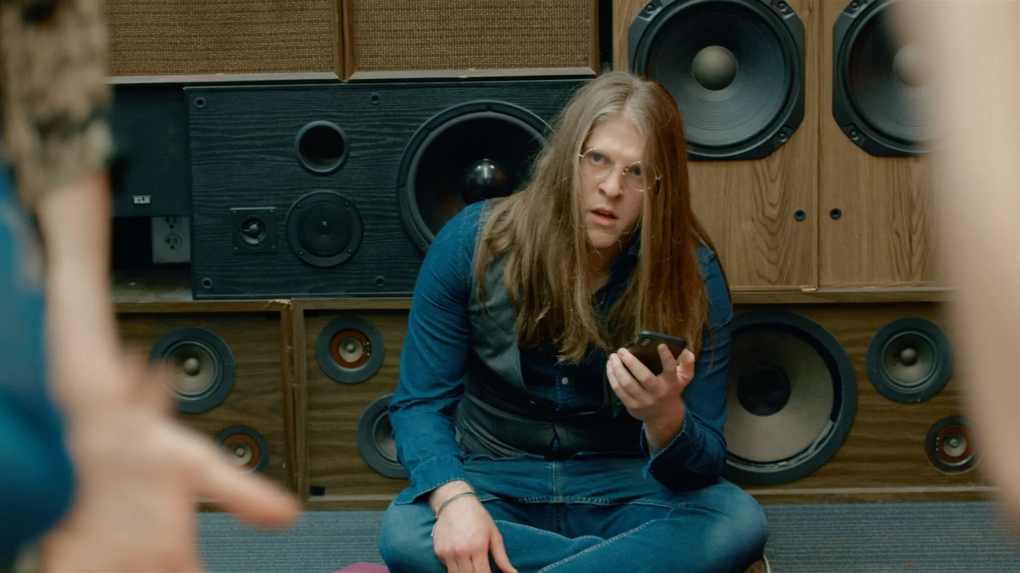 A young woman with long hair, glasses, and wearing a blue shirt sitting on the floor in front of large speakers, holding a phone and looking surprised or confused.