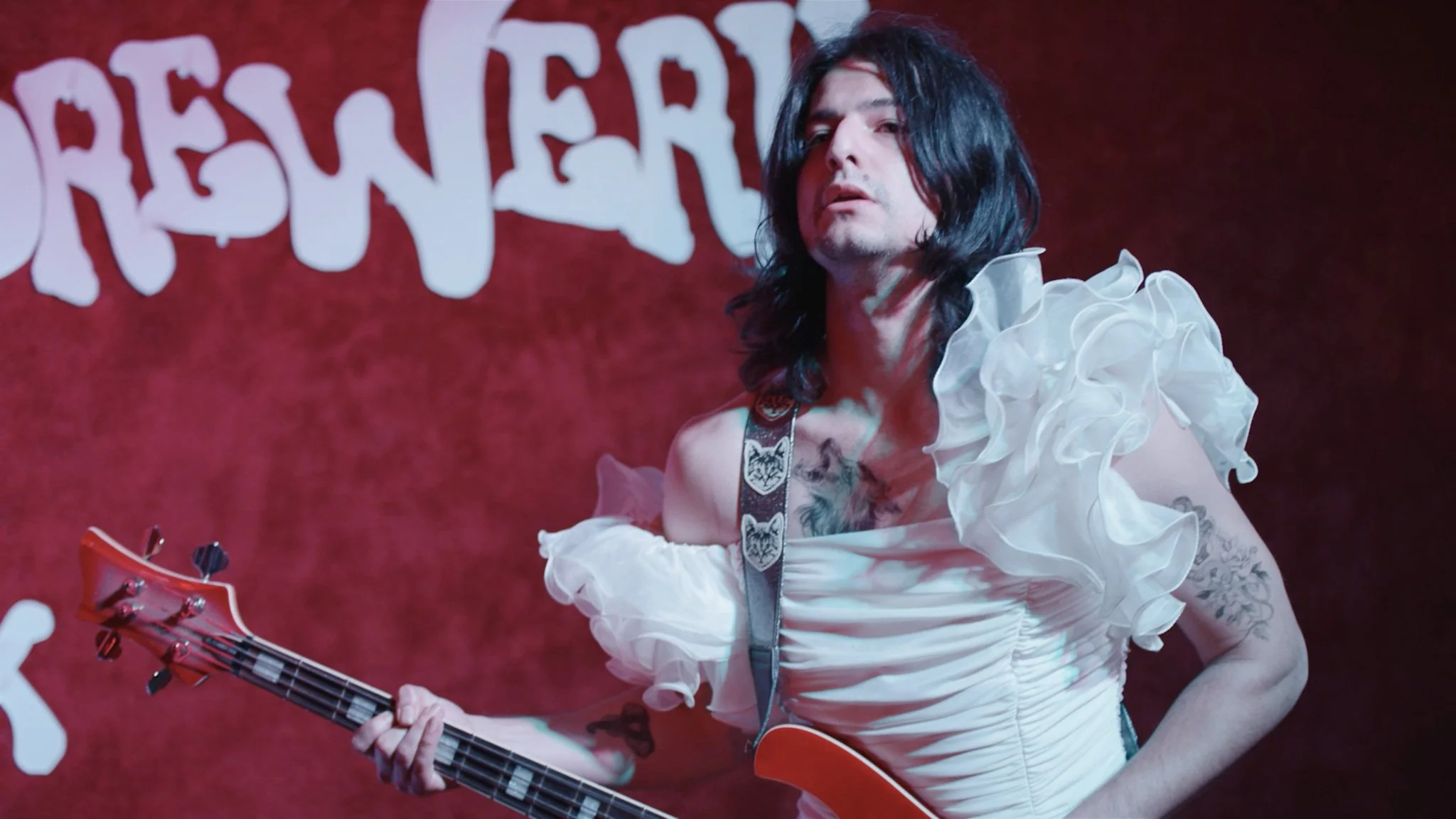 A person with long black hair and tattoos, wearing a white ruffled top, holding a guitar on stage with a red background and partially visible white text.