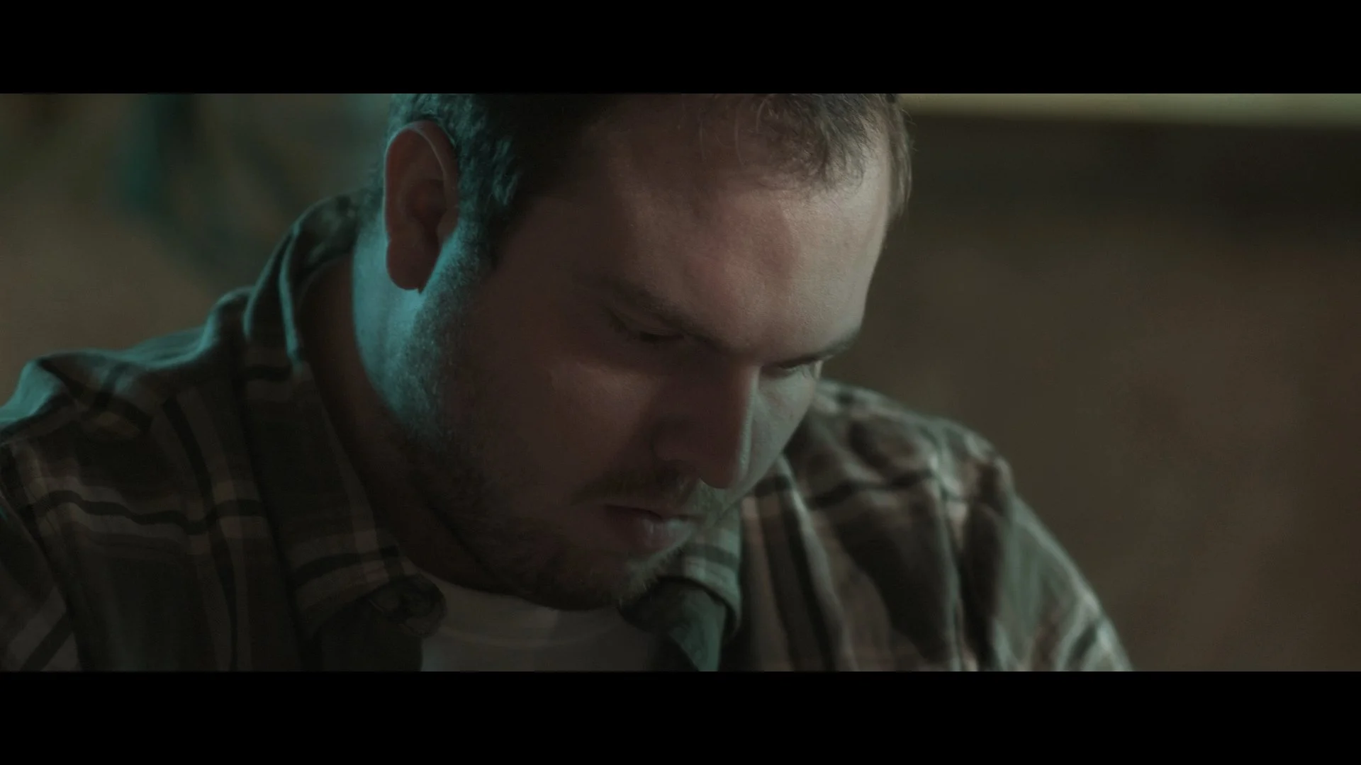 A man with short hair and a beard looking downward, wearing a checkered shirt, in a dimly lit room.