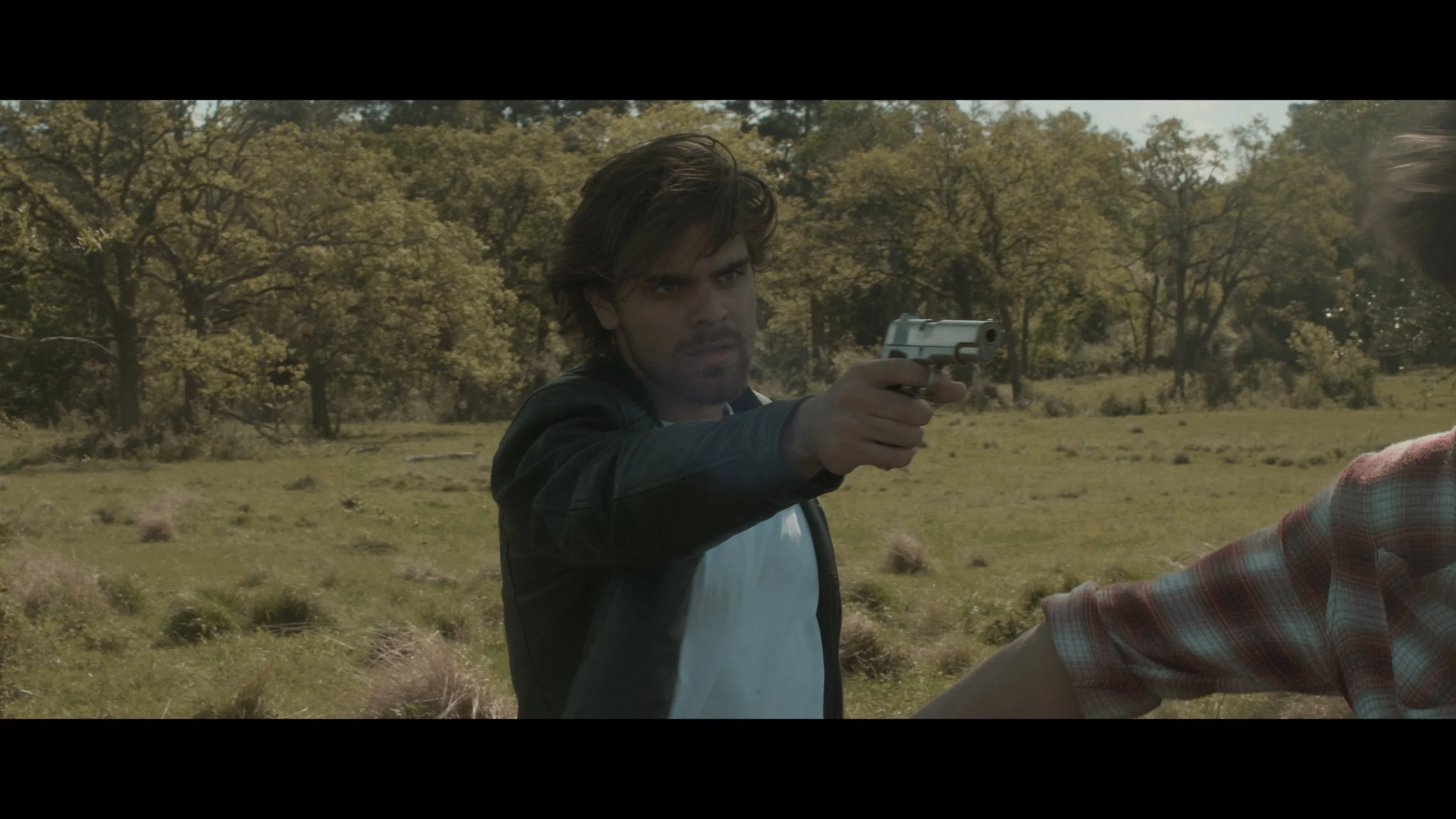 A man with dark hair and a serious expression pointing a handgun at someone outside the frame in a grassy field with trees in the background.