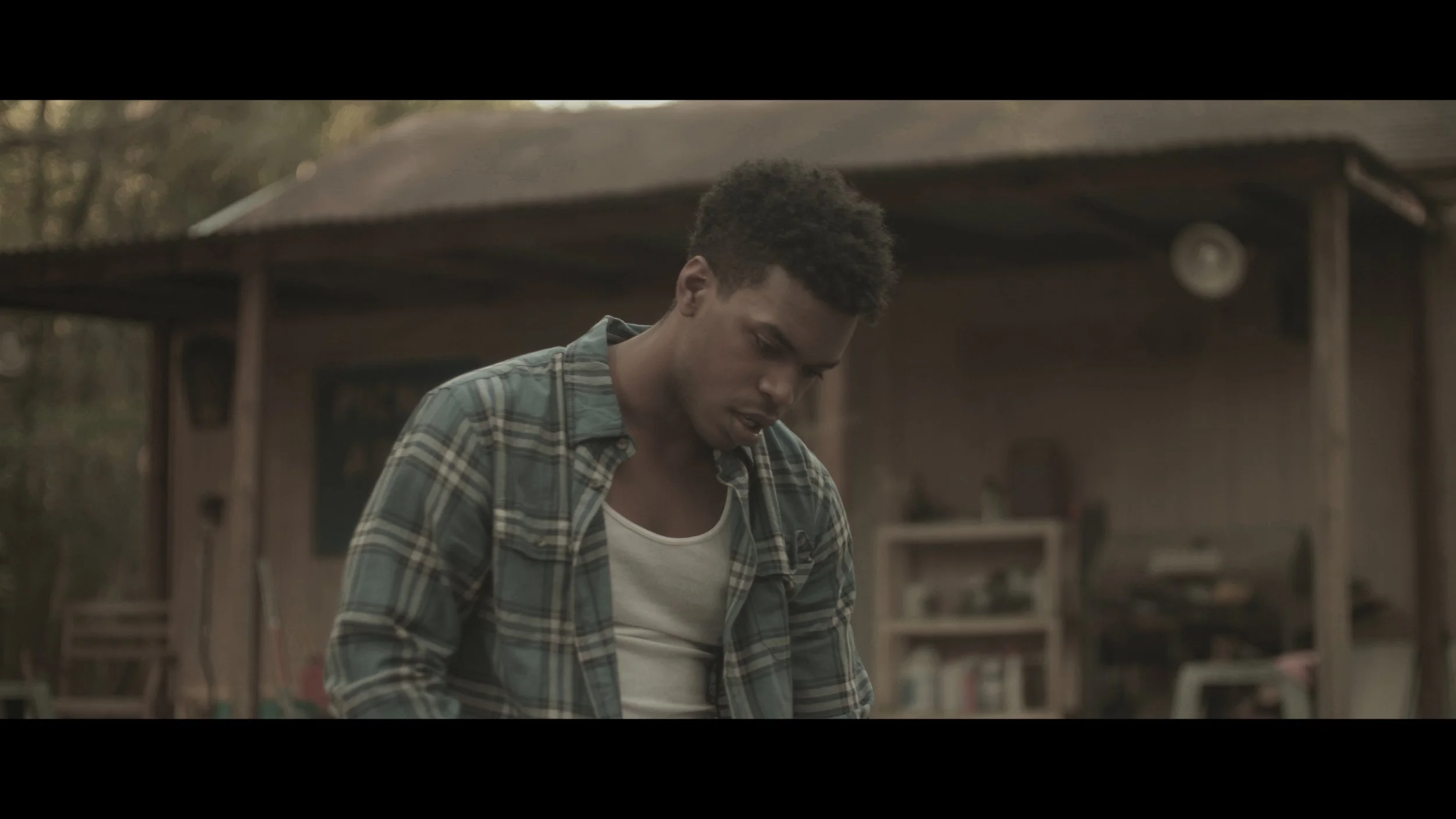 A young man with short curly hair wearing a plaid shirt over a white tank top looking down outdoors in a rustic setting.