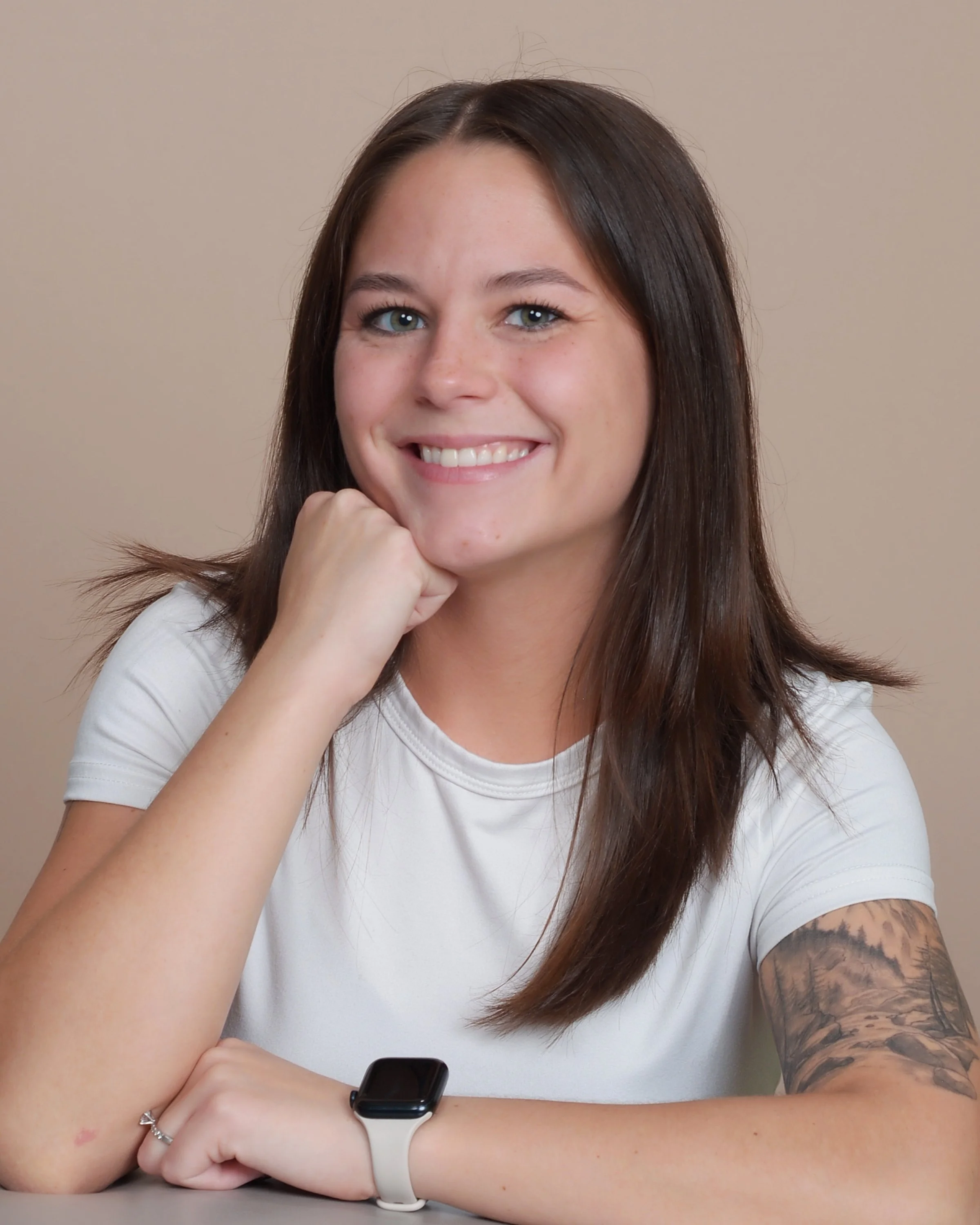 A young woman with long brown hair, blue eyes, and light skin is smiling while resting her chin on her hand. She is wearing a white t-shirt and a smartwatch on her left wrist. She is sitting at a table with a beige background. Her right arm displays a tattoo of a landscape scene, and she wears a silver ring on her left ring finger.