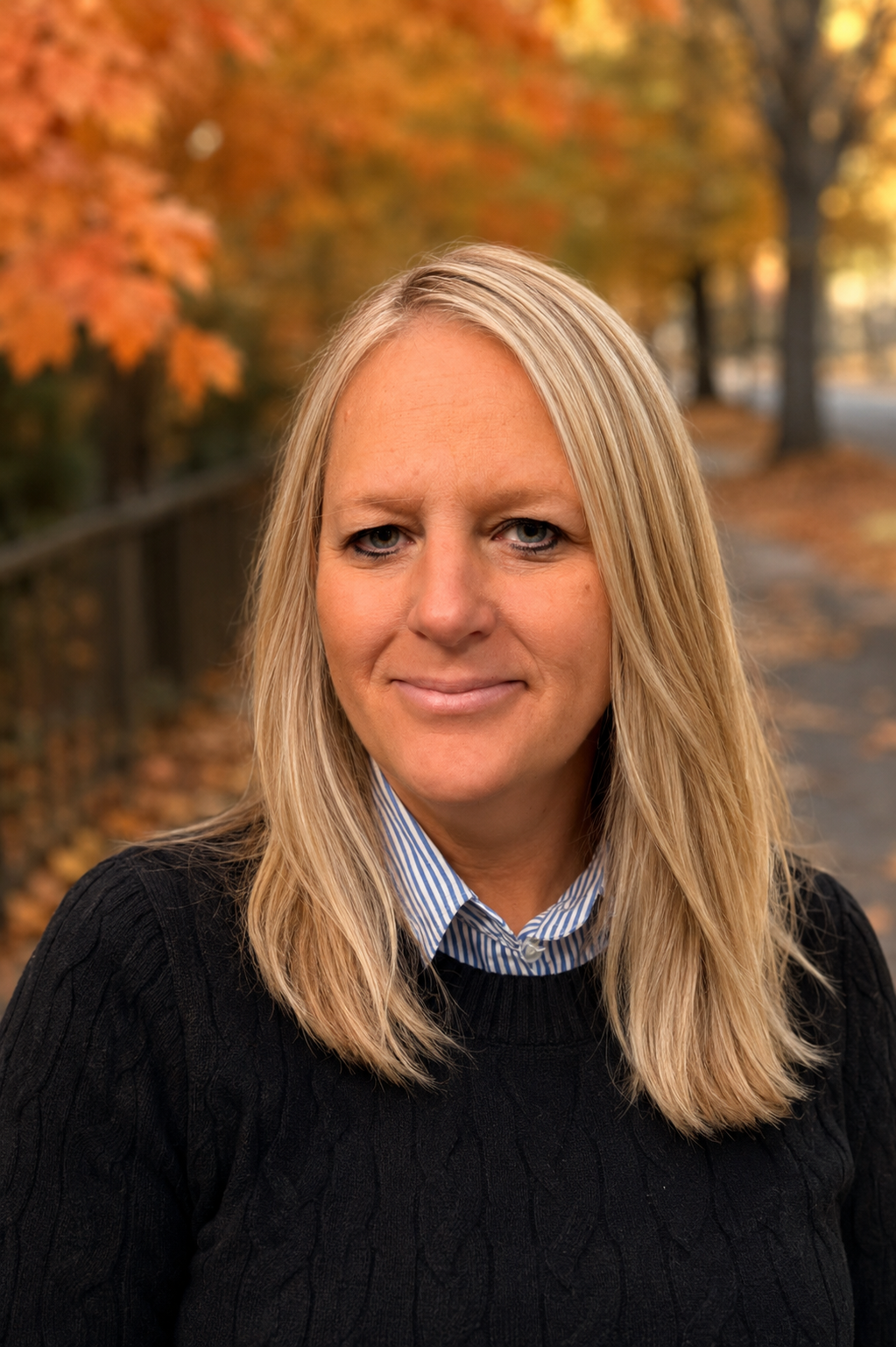 A woman with blonde hair and blue eyes smiling outdoors in autumn, with colorful fall leaves on trees and a wooden fence in the background.