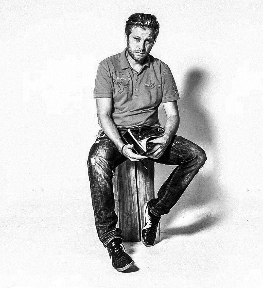 Black and white photo of a man with styled hair and beard sitting on a wooden box, holding a smartphone, against a plain background.