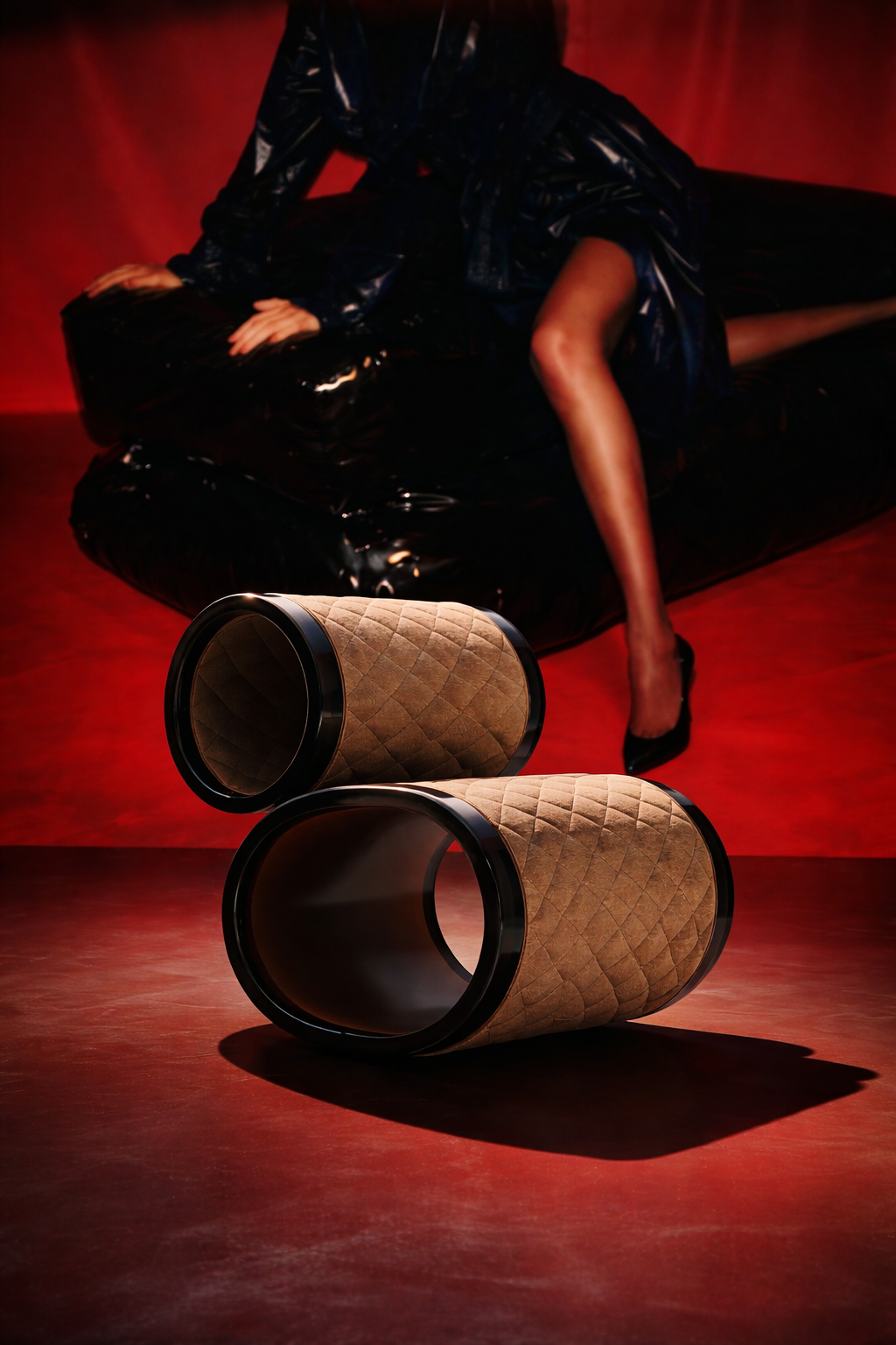 Close-up of two cylindrical objects with quilted beige fabric and black rims, placed on a red floor. In the background, a woman dressed in black with shiny material and high heels is sitting on a black leather couch with a red backdrop.