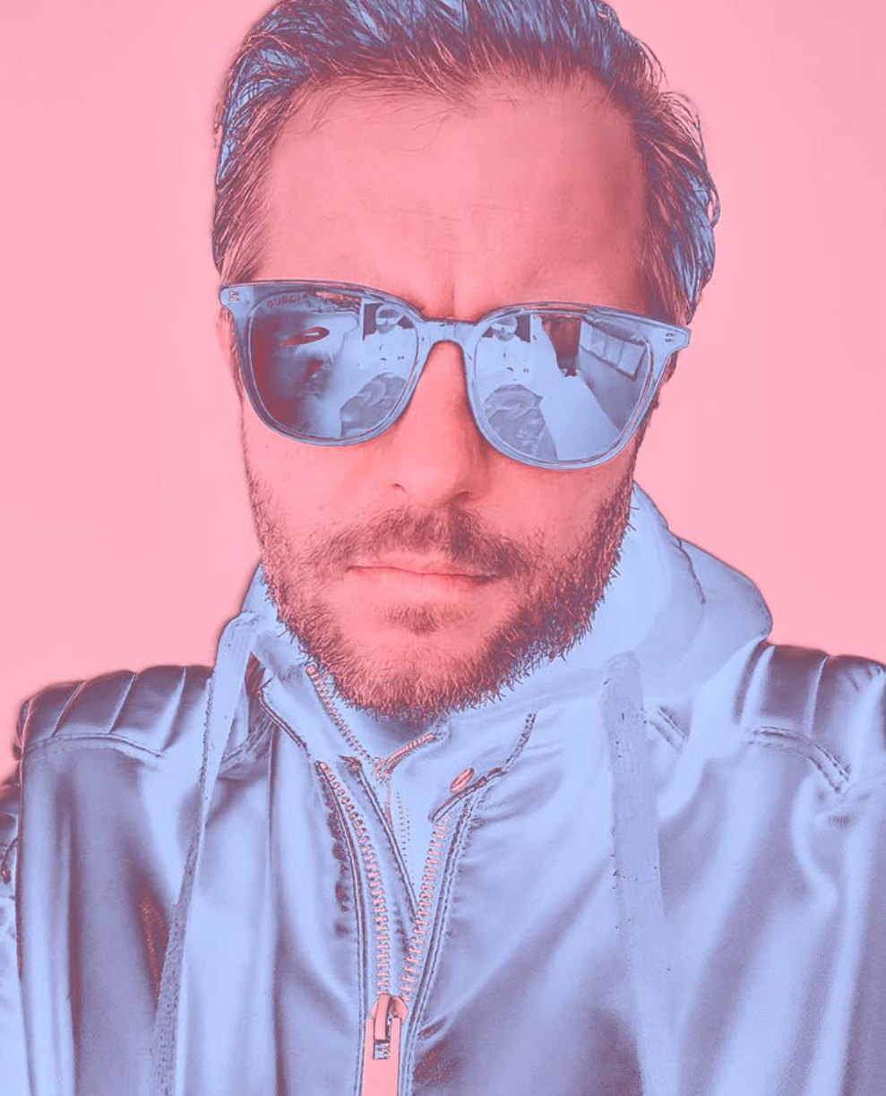 A man with sunglasses and a light-colored jacket poses against a pink background.