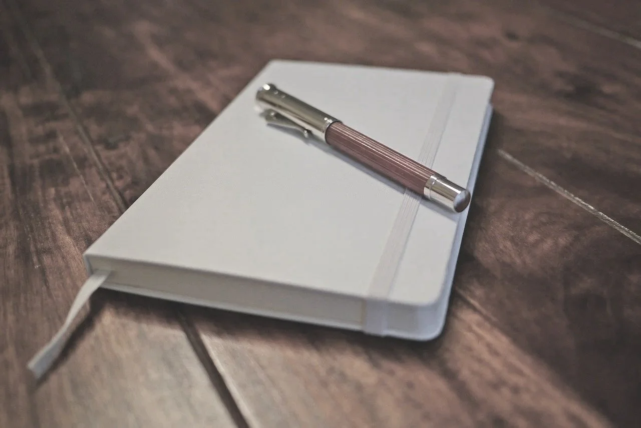 A white hardcover notebook with an elastic band, a white ribbon bookmark, and a pink and silver pen resting on top, placed on a wooden surface.
