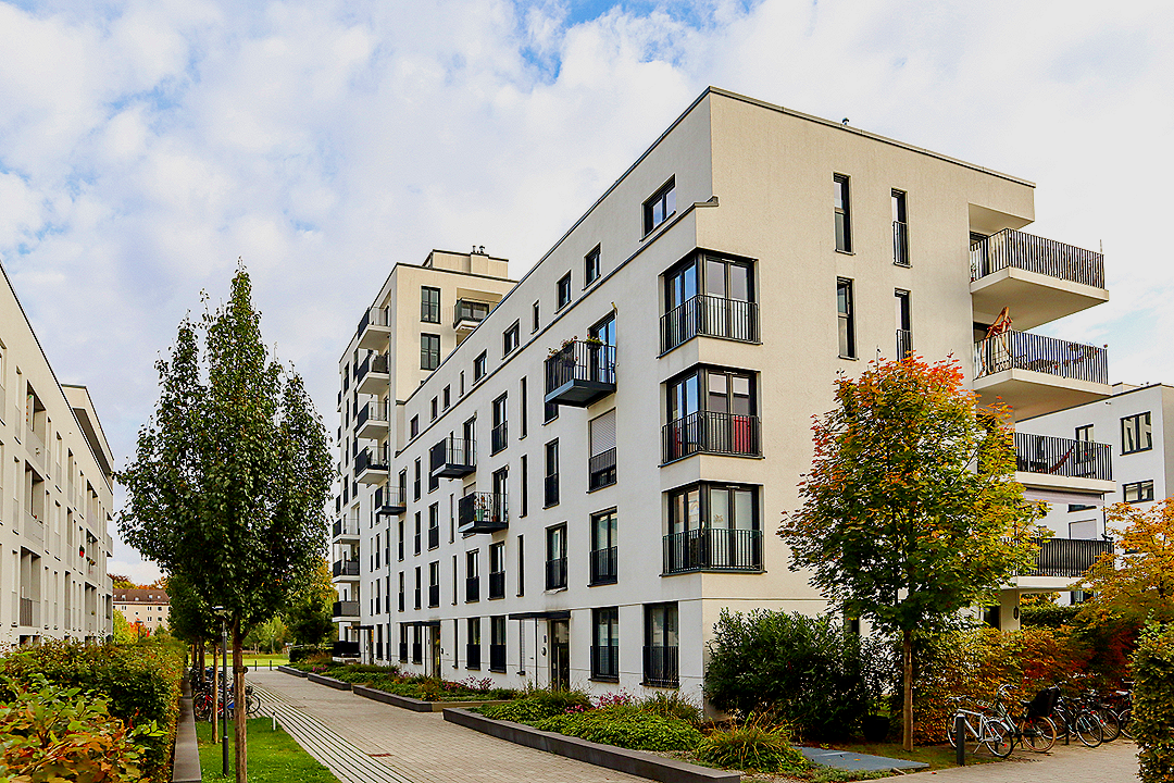 Modern white apartment building surrounded by trees and a paved walkway.
