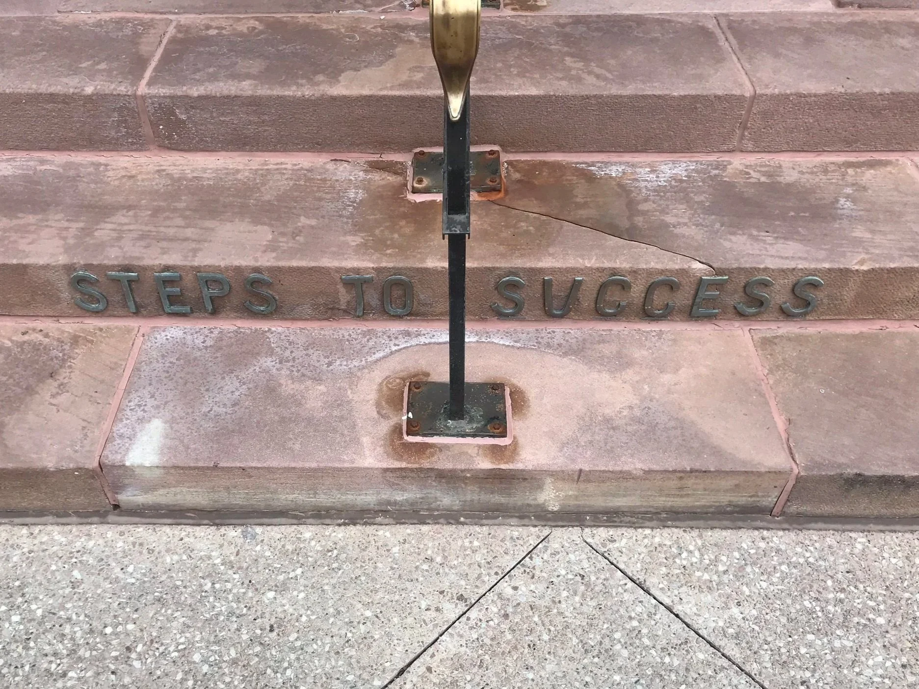 The image shows a set of stone steps with the words 'STEPS TO SUCCESS' embedded in the front edge of the bottom step, and a metal railing with a mounting base in the center.