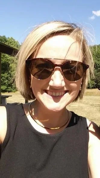 A smiling woman wearing sunglasses and a black sleeveless top outdoors on a sunny day.