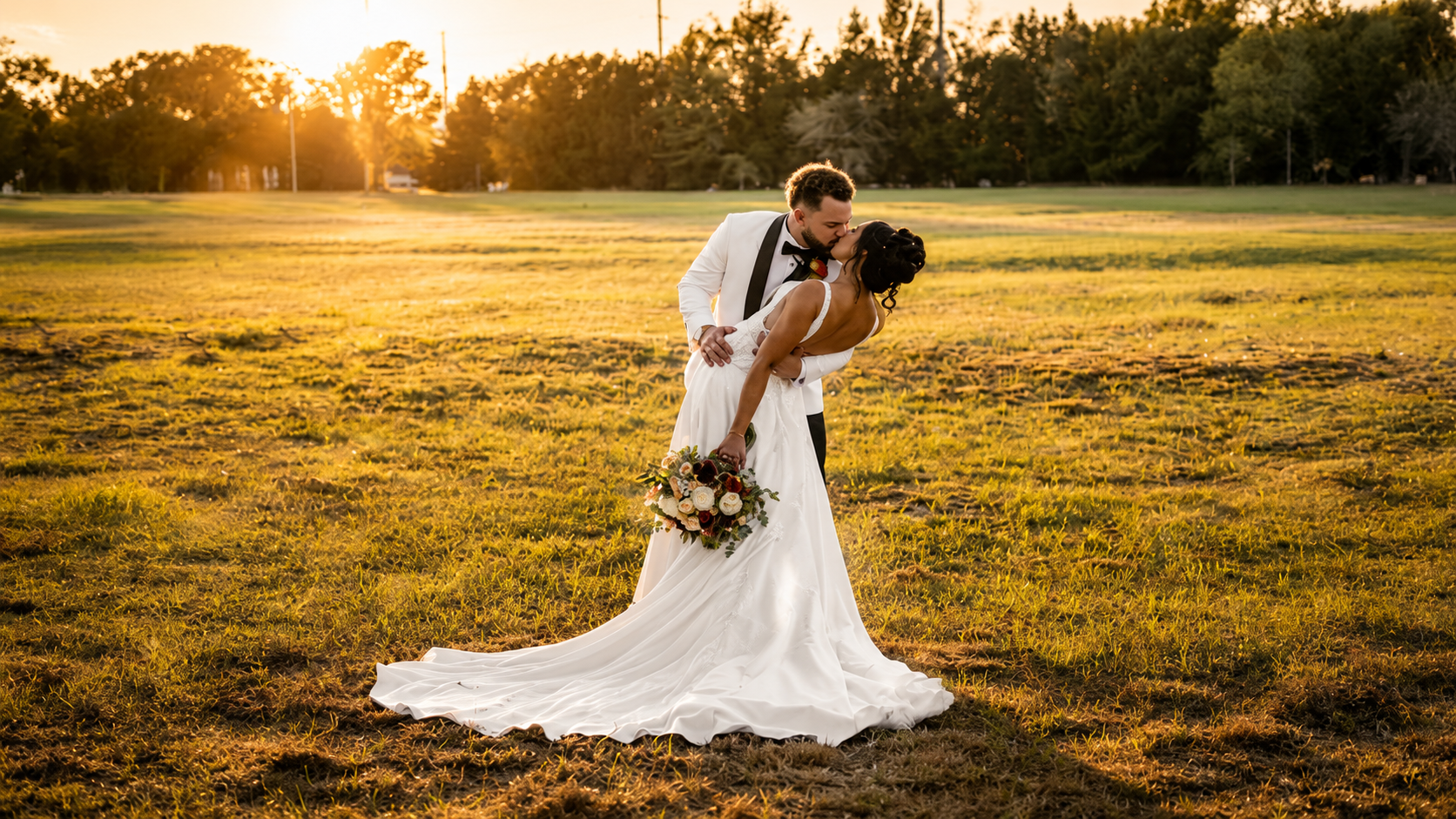 A bride and groom sharing a kiss during sunset in an open grassy field, with the bride holding a bouquet of flowers.