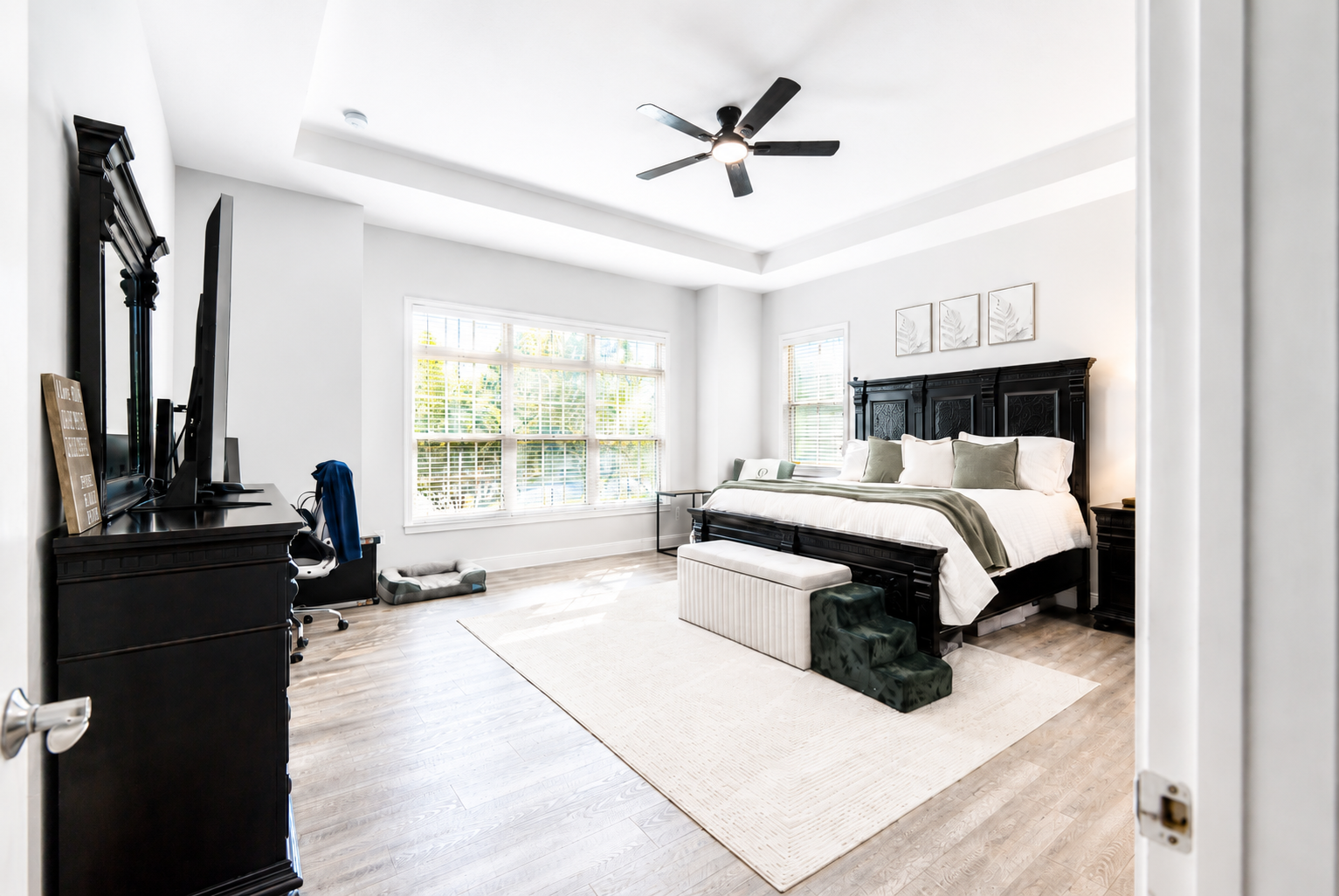 A spacious bedroom with a large bed, black wooden headboard, white bedding, decorative pillows, and a bench at the foot of the bed. There is a ceiling fan, two side tables, framed wall art, and big windows with blinds letting in natural light. A desk with a monitor, chair, and some items is visible on the left.