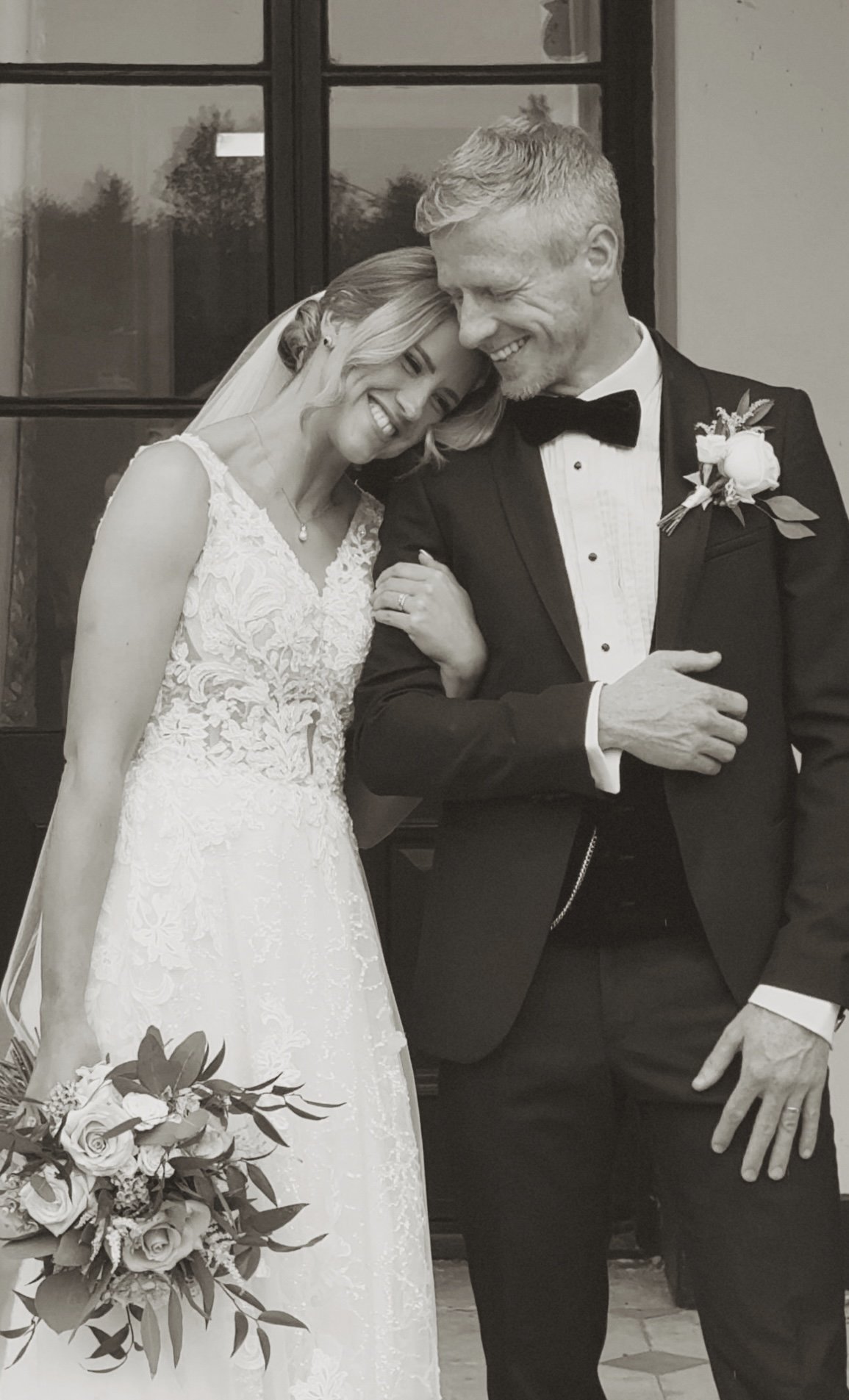 Black and white photo of a bride and groom at their wedding, standing close together in front of a window, with the bride holding a bouquet of flowers.