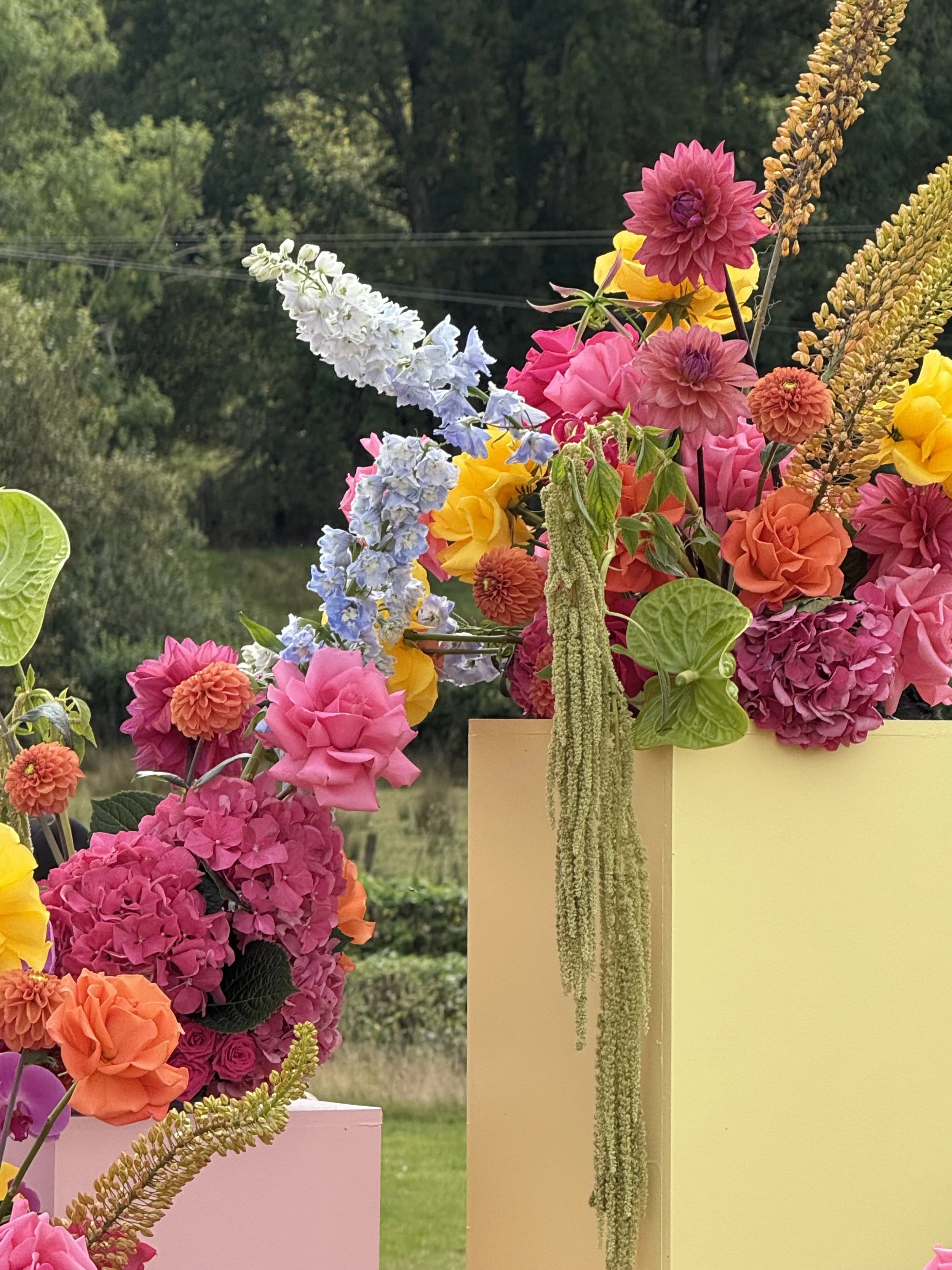 Colourful flower arrangement with pink, orange, yellow, purple and blue flowers in a yellow vase outdoors, with greenery in the background.