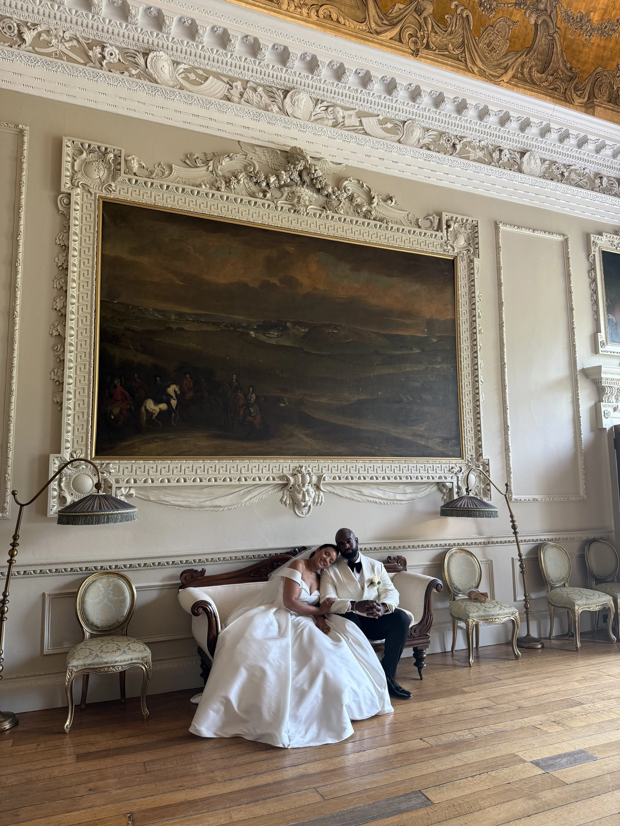 A bride and groom sitting on a vintage sofa in an ornate, historic room with classical decor, large painting, and four chairs along the wall.