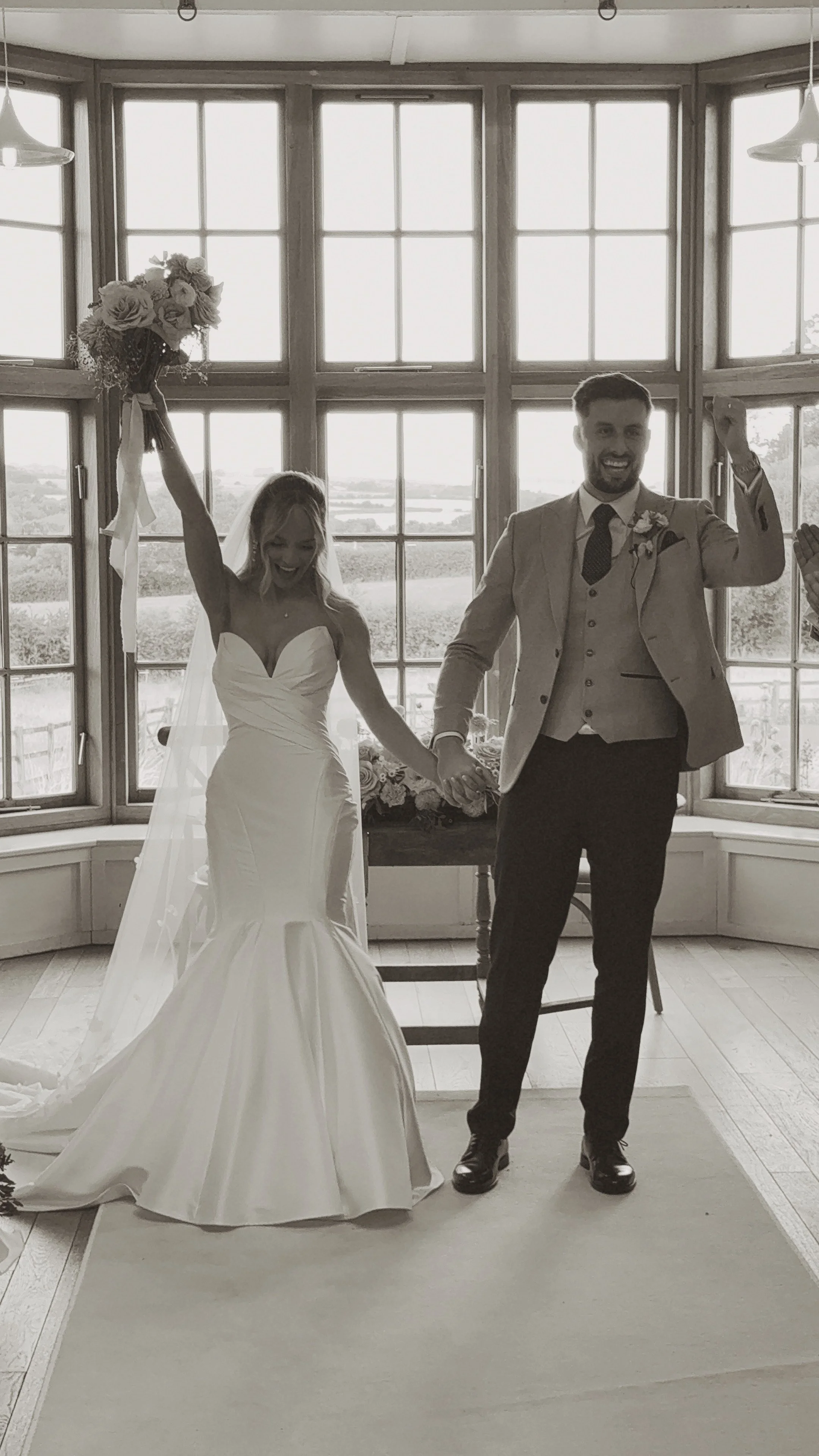 A bride and groom celebrating as their ceremony is coming to an end. They are holding hands in the air and smiling, with a large window in the background.