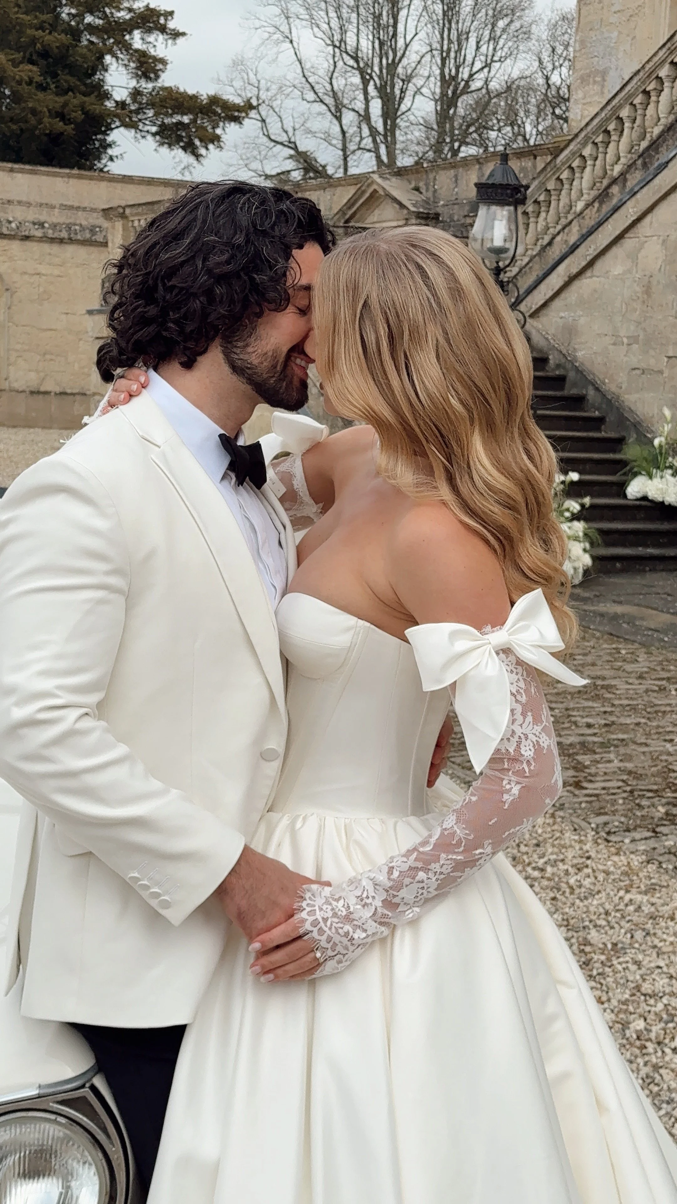 A bride and groom share a close, intimate moment outdoors on their wedding day, with stone stairs and greenery in the background.