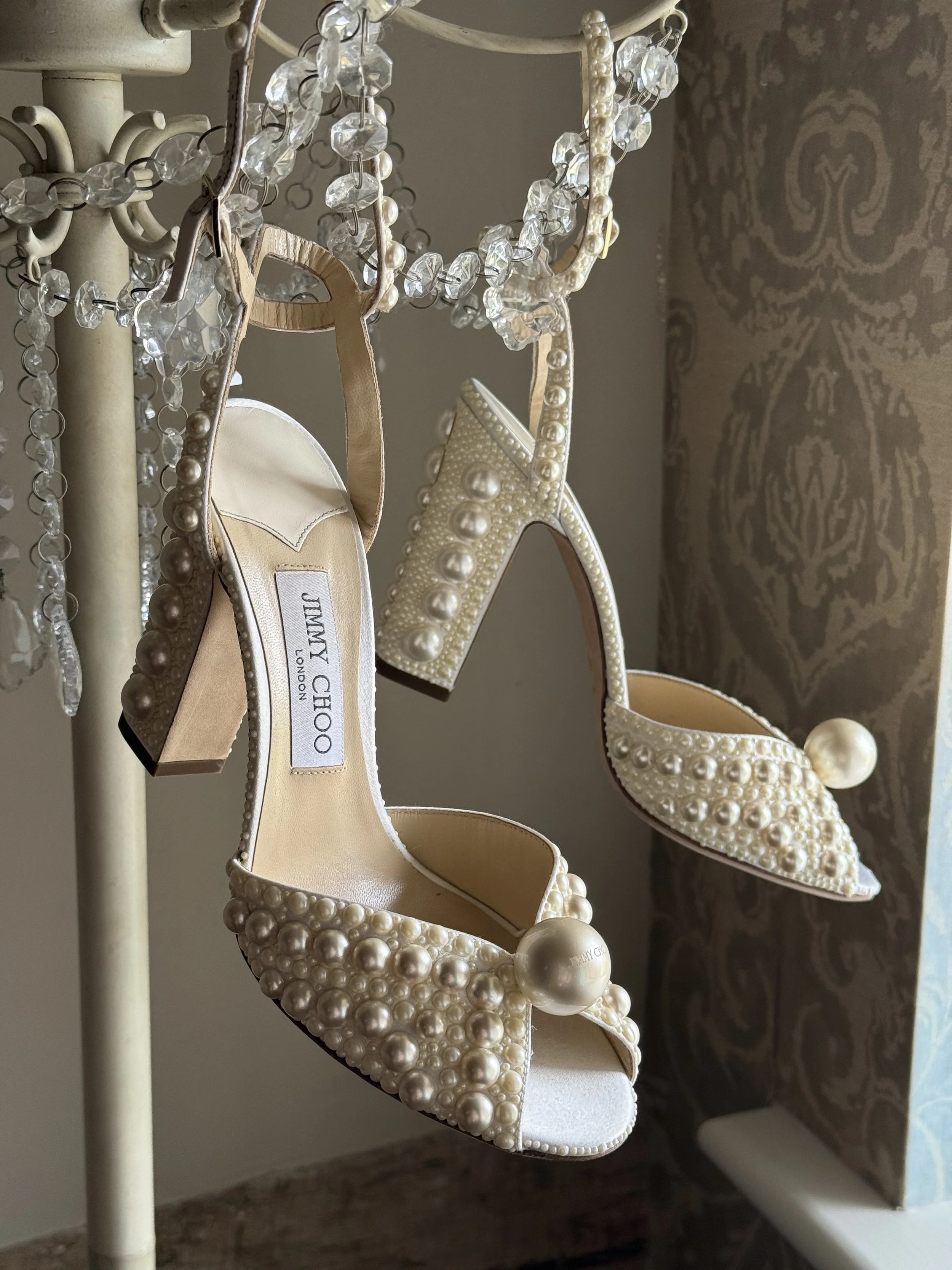 A pair of high heel shoes decorated with pearls and featuring large pearl accents on the toe, hanging on a vintage chandelier with crystal beads, in a room with patterned wallpaper.