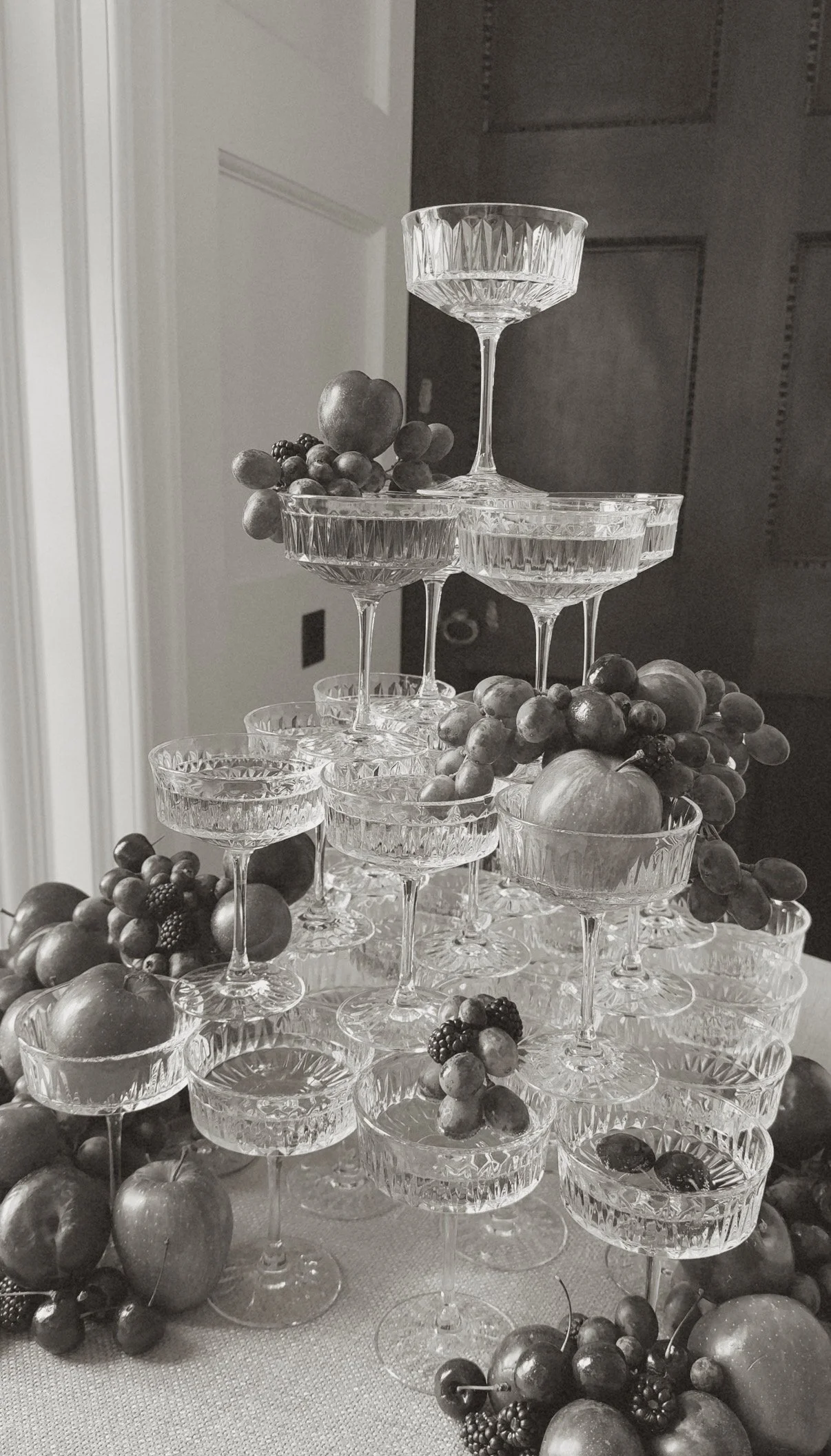 A pyramid of glass dessert cups arranged with grapes and apples, decorated with berries, on a table with additional fruits.
