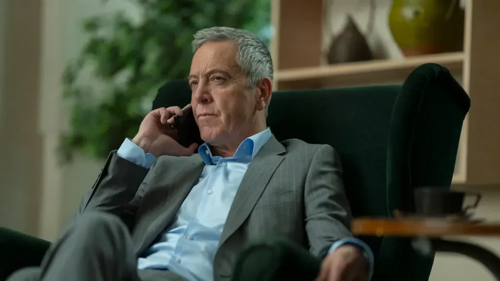 A man in a gray suit sitting in a black armchair, talking on a cellphone, with a serious expression in a professional office setting.