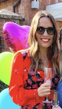 A smiling woman wearing sunglasses and a red floral dress, holding a glass of champagne, standing in front of colorful balloons outdoors.