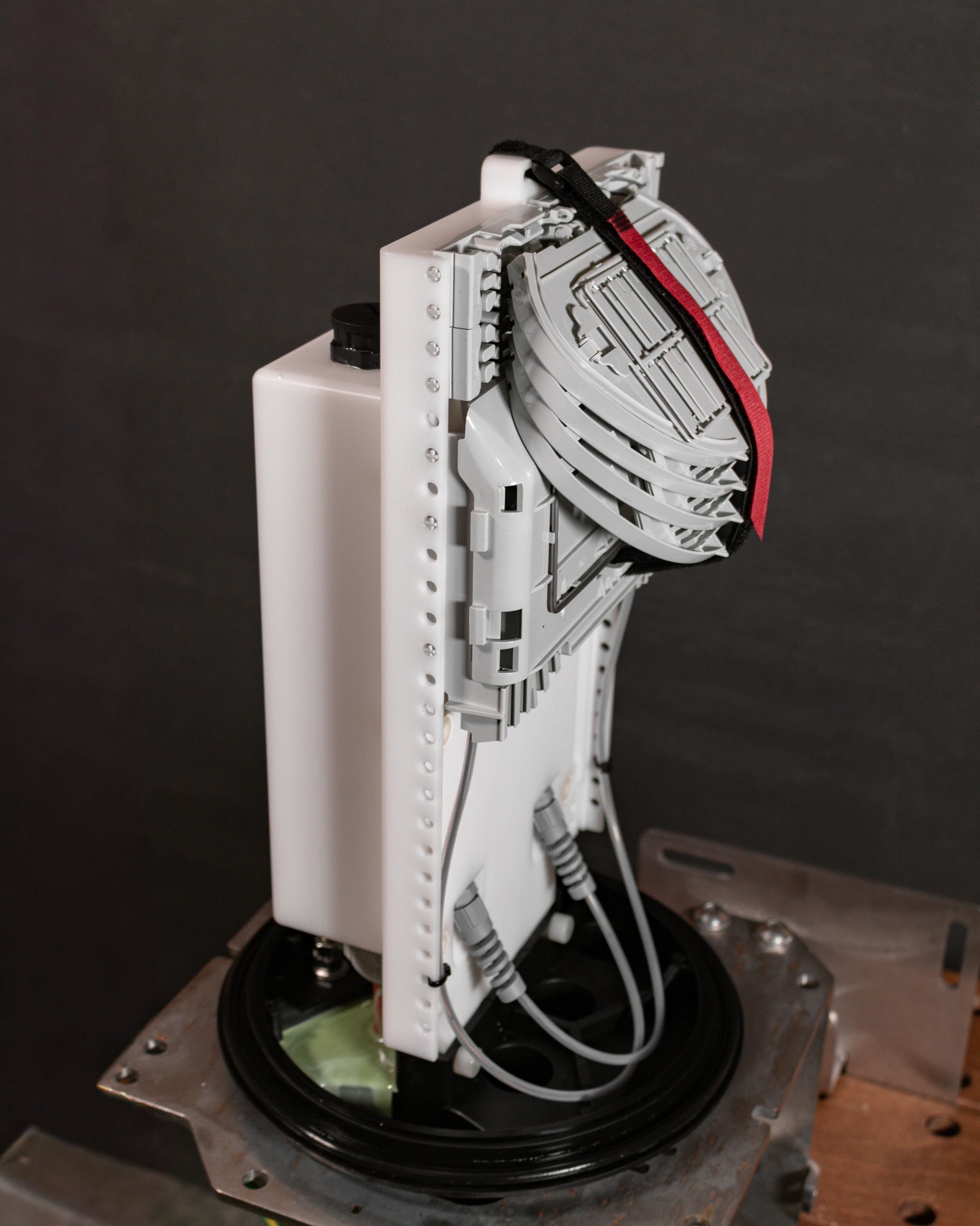 A close-up view of a partially disassembled robotic or mechanical device with gray and white components, wires, and a black and red strap, set against a dark background.