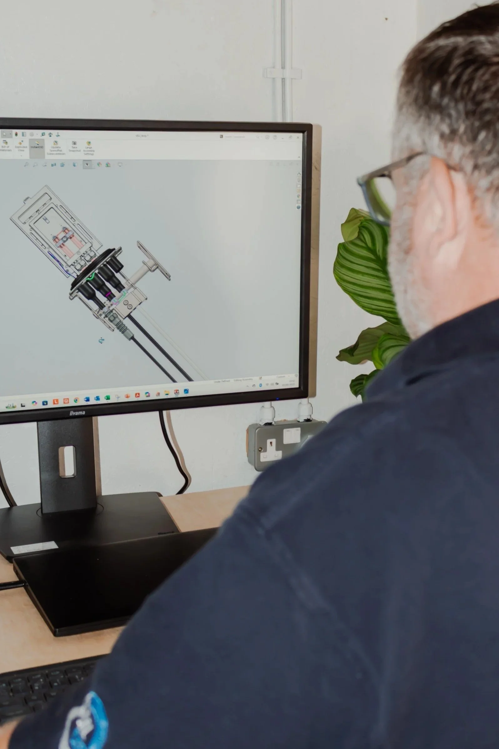 A man sitting at a desk, looking at a computer monitor displaying a technical CAD drawing of a mechanical device.
