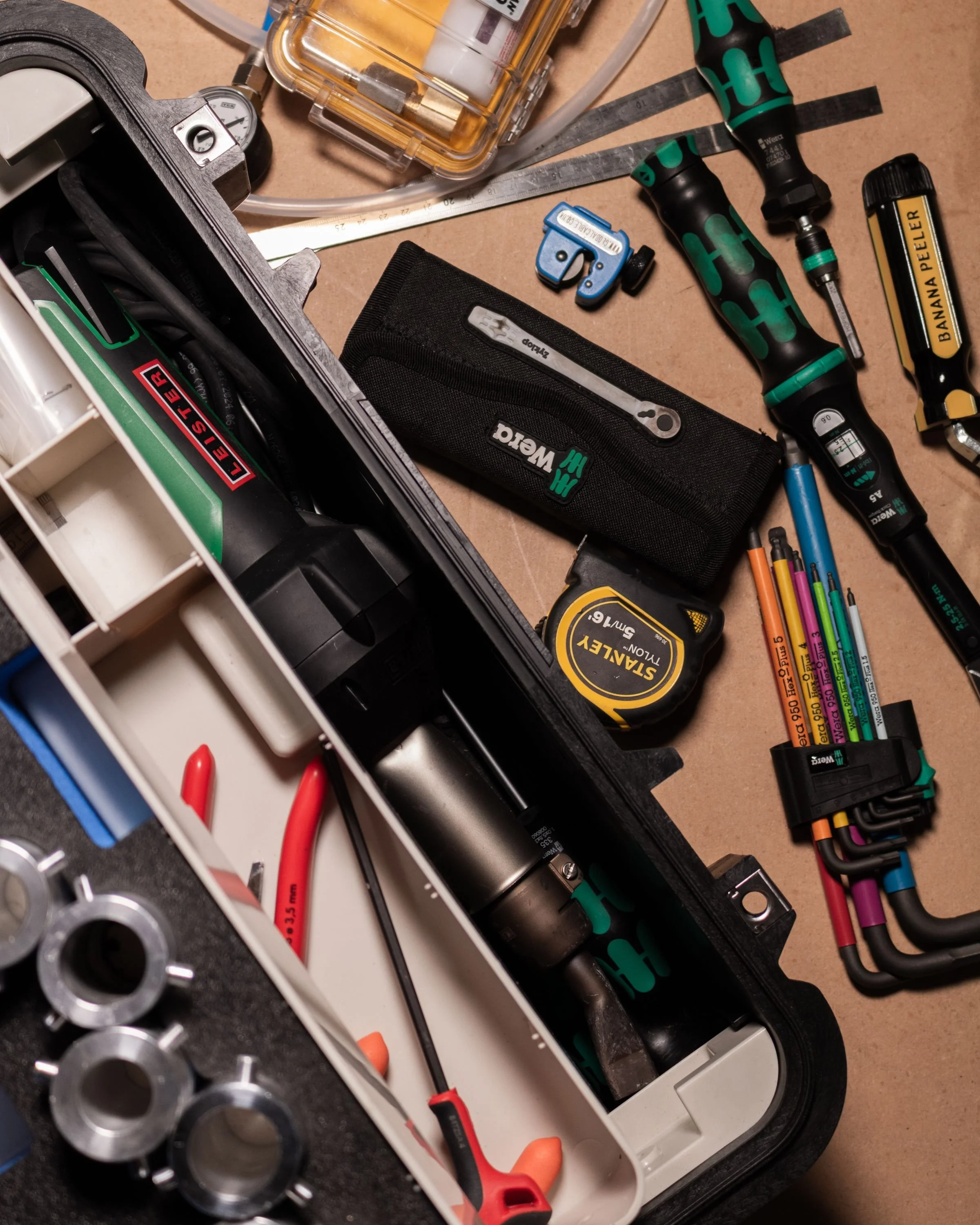 A cluttered workbench with various tools and equipment, including screwdrivers, a tape measure, colored pencils in a holder, a small black case, a power drill, and metal pipe fittings.