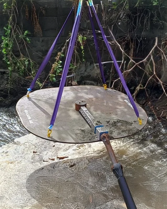 A large round mirror positioned in a river, held up by purple straps attached to a tripod structure. A trowel or scraper tool is resting on the mirror, possibly for cleaning or adjusting.