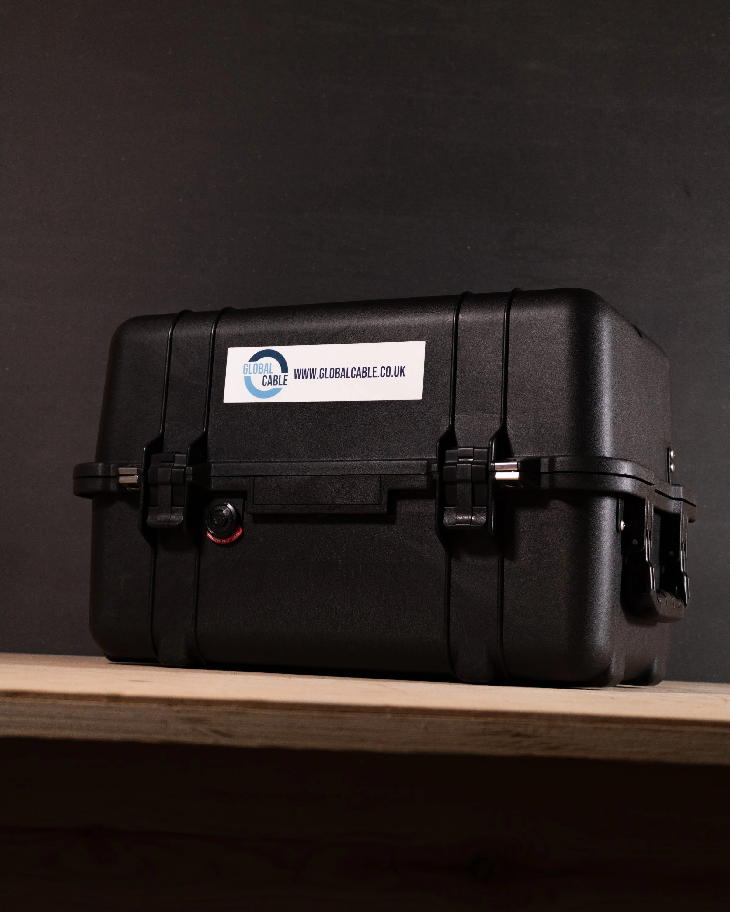 Black protective carrying case with a label for Global Cable and a website URL, placed on a wooden shelf against a dark background.