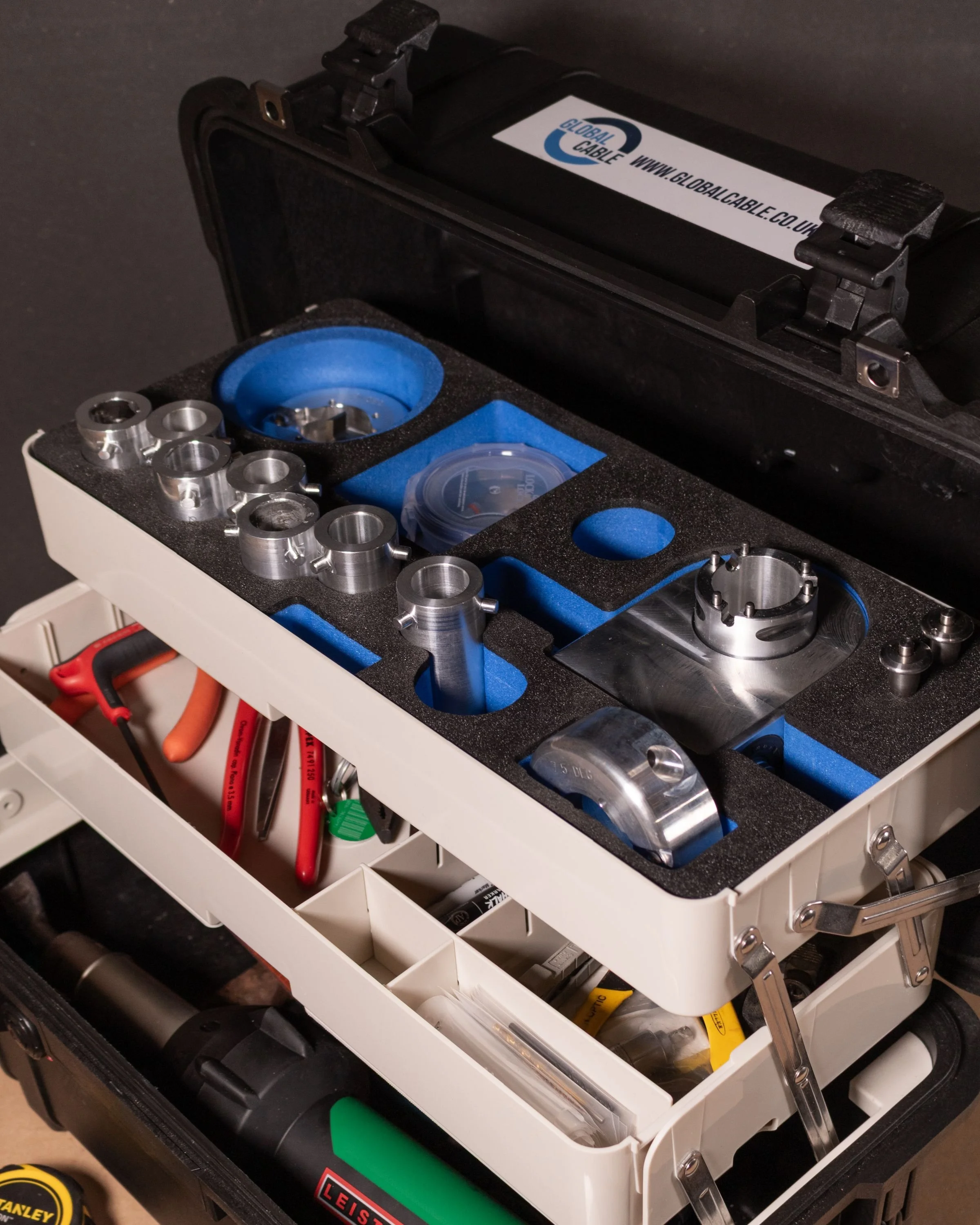 A toolkit with various metallic parts, tools, and accessories organized in foam and plastic compartments, including a black case, red-handled pliers, a flashlight, and other small components.