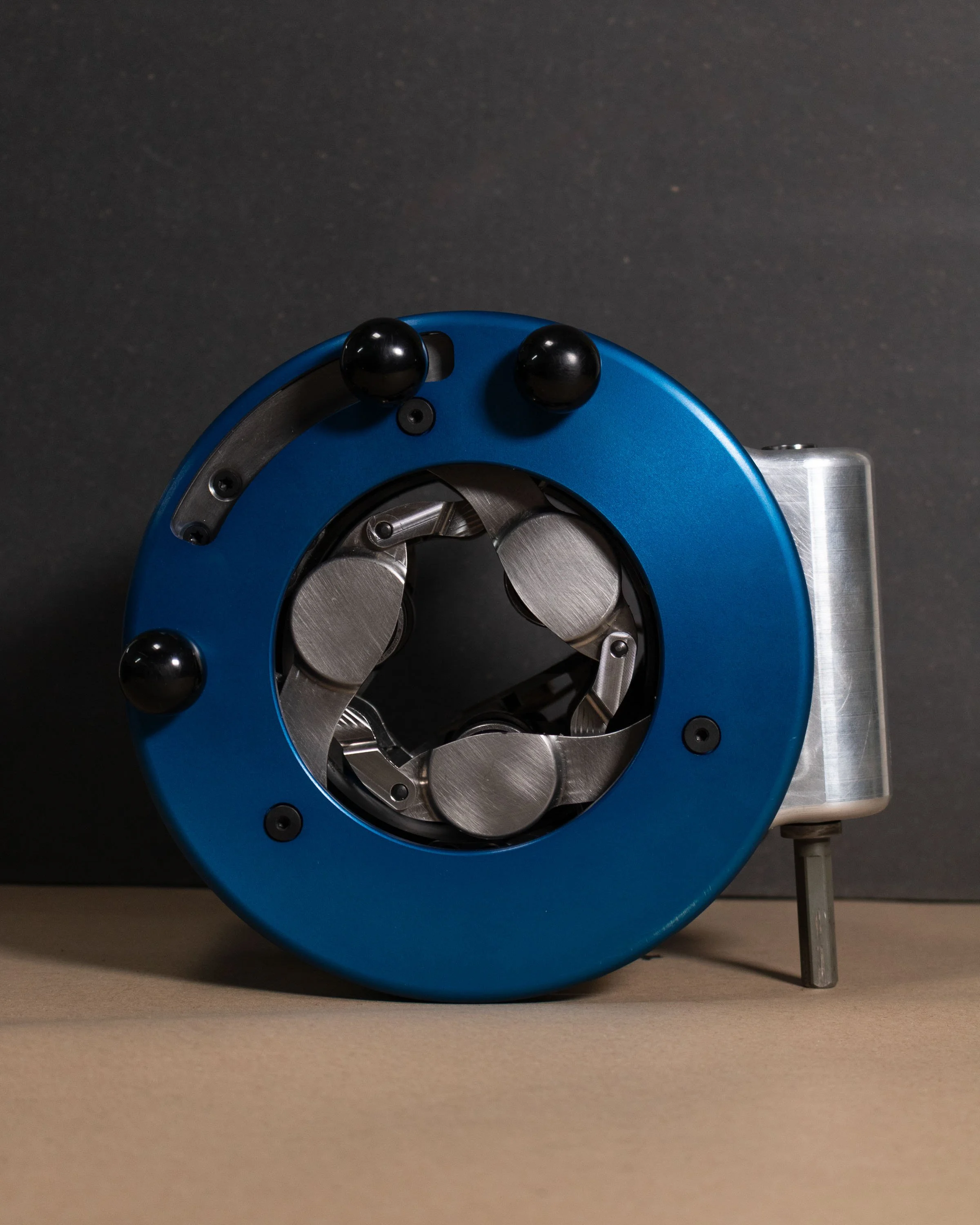 A mechanical device with a blue circular outer housing, black spherical handles, and metallic internal components visible through a central opening, set against a dark background.