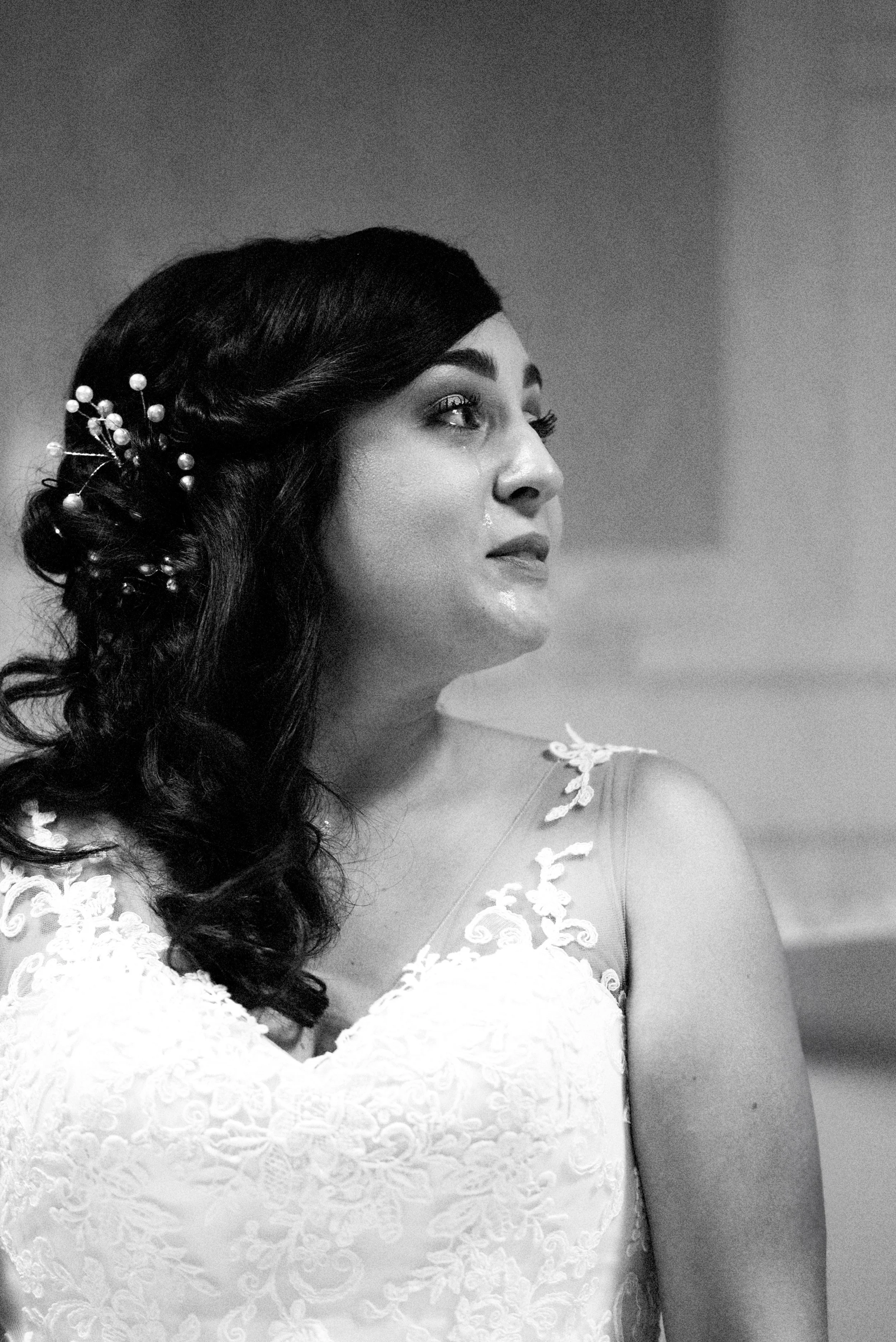 Black and white portrait of a woman with dark hair styled with decorative hairpins, wearing a lace dress, looking to her right with a tear on her cheek.