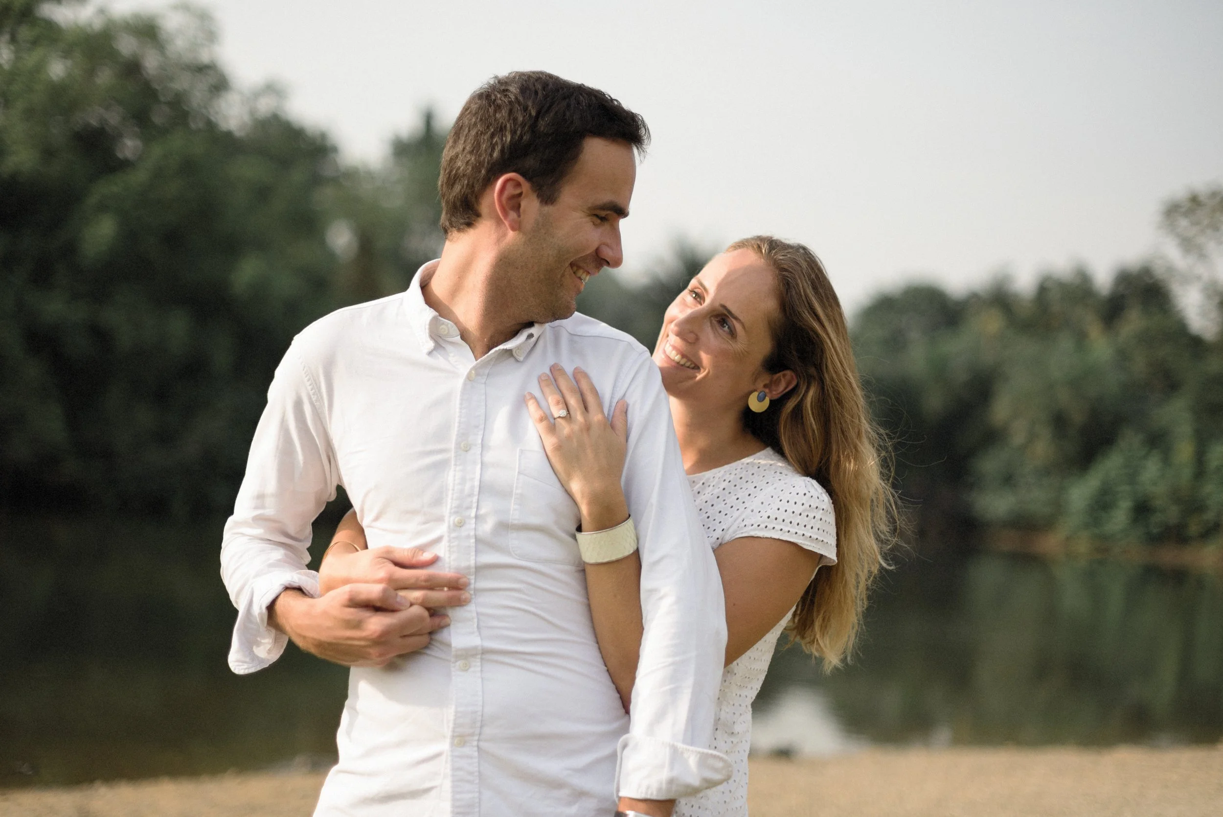 A happy couple smiling and looking at each other outdoors near a body of water with trees in the background.