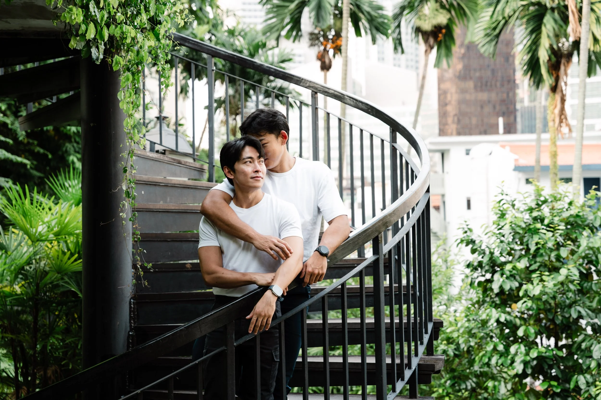Two men standing on a spiral staircase outdoors, surrounded by green plants and trees, with a cityscape in the background, one man embracing the other from behind.
