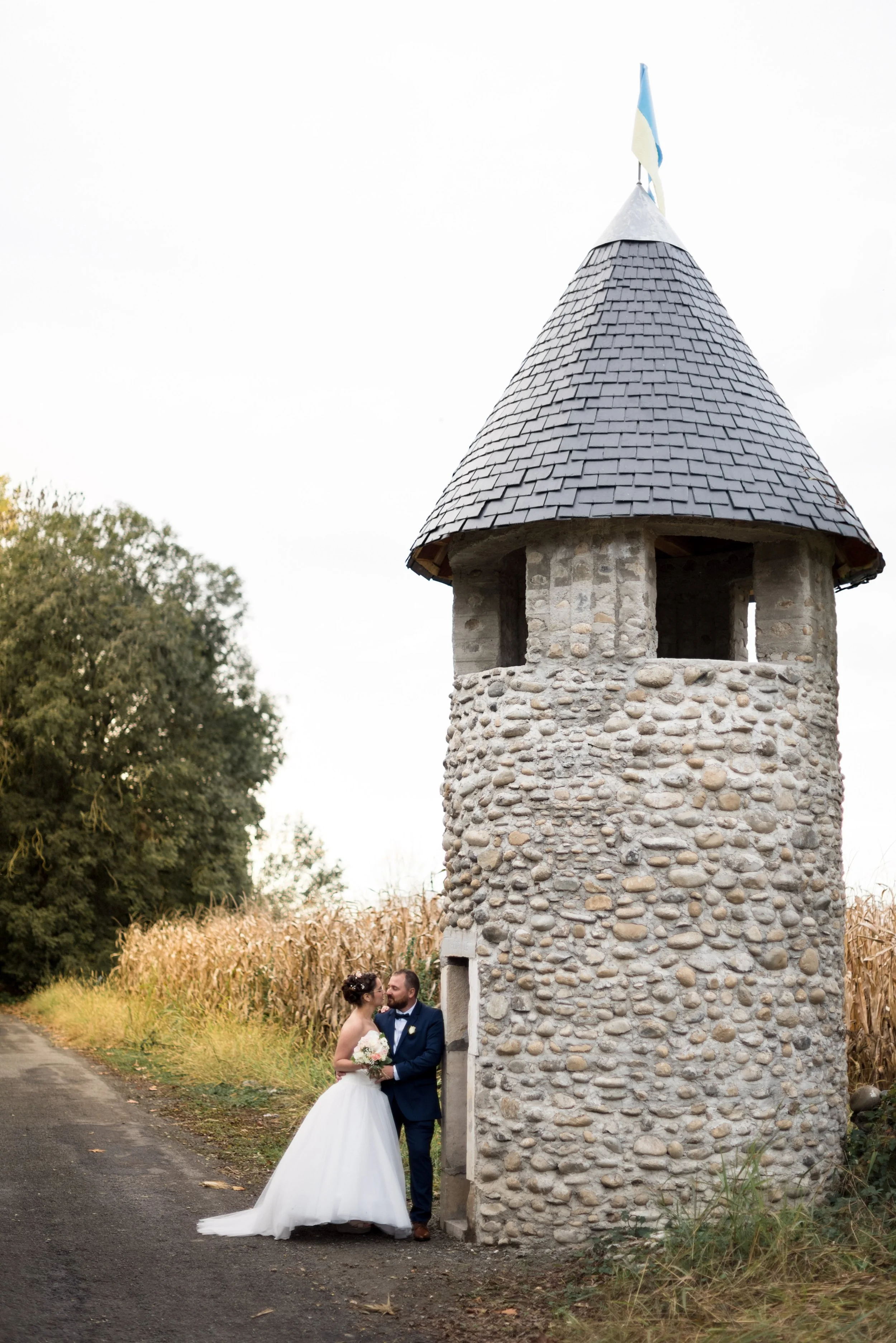 A bride and groom standing together beneath a small stone tower with a slate roof, outdoors near a field of corn.